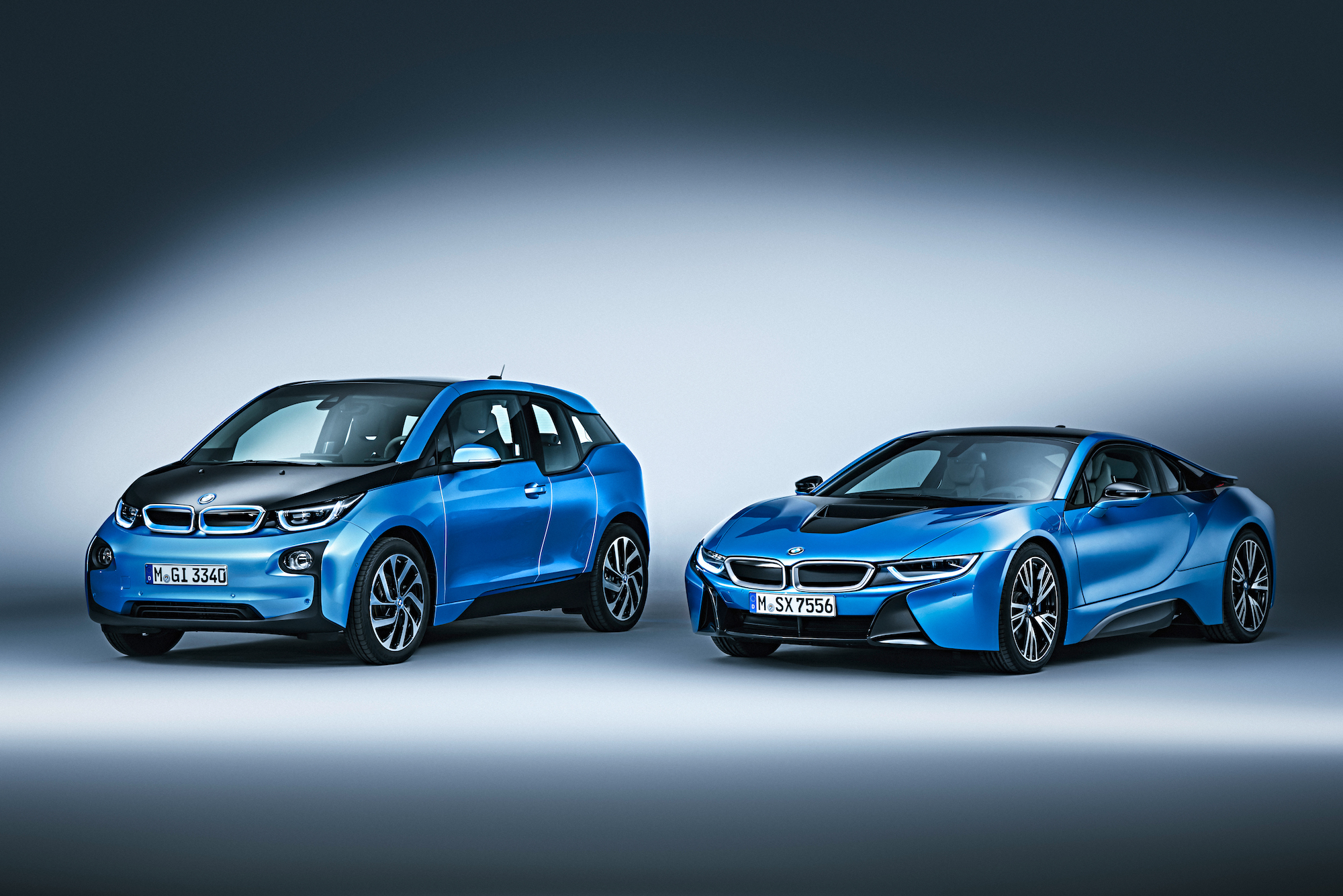 Zwei blaue BMW-Modelle, links ein kompakter i3 und rechts ein sportlicher i8, stehen nebeneinander auf grauem Hintergrund.