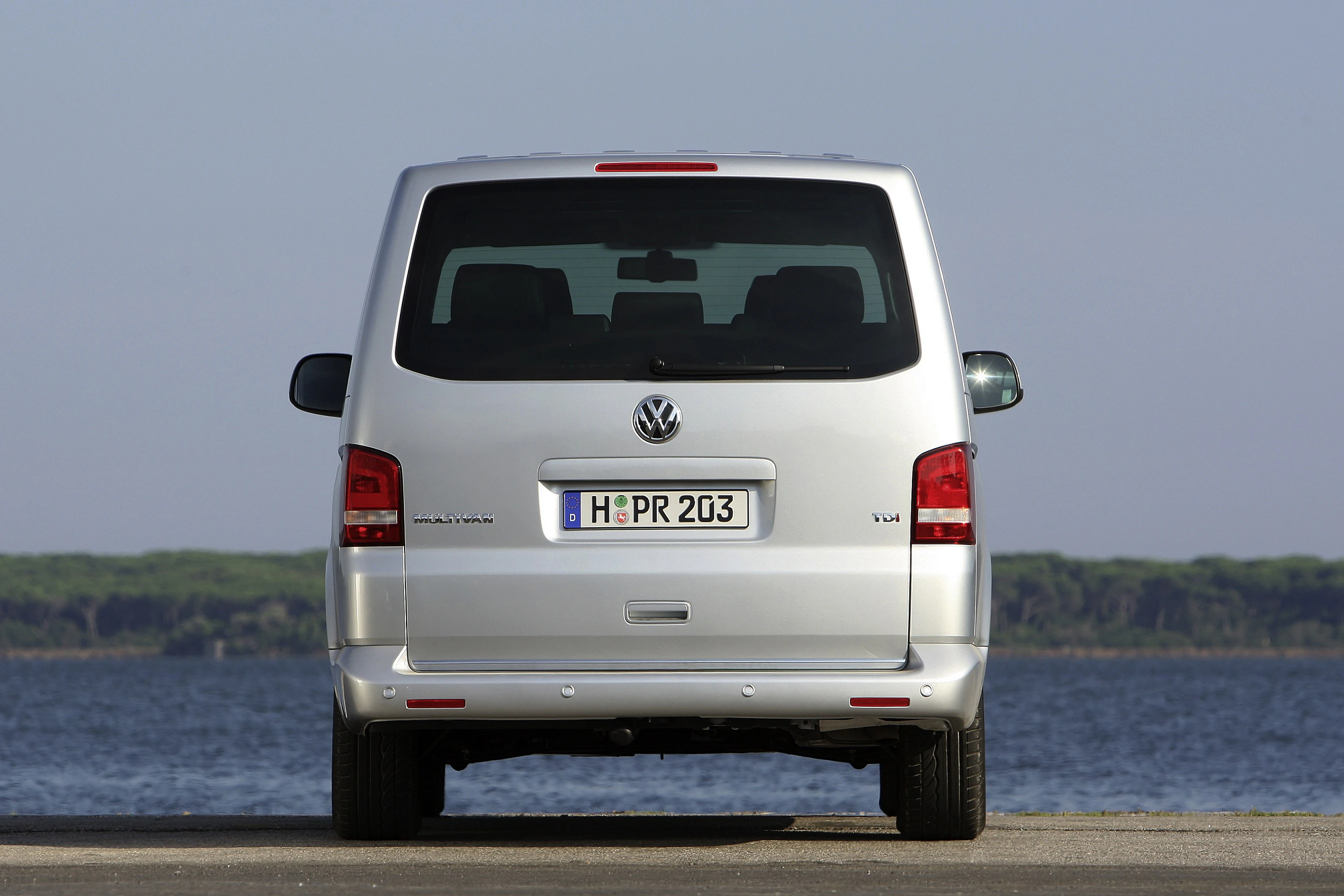 Silberner Volkswagen Multivan, Heckansicht, mit deutschem Kennzeichen, geparkt vor einem See mit bewaldetem Ufer im Hintergrund.