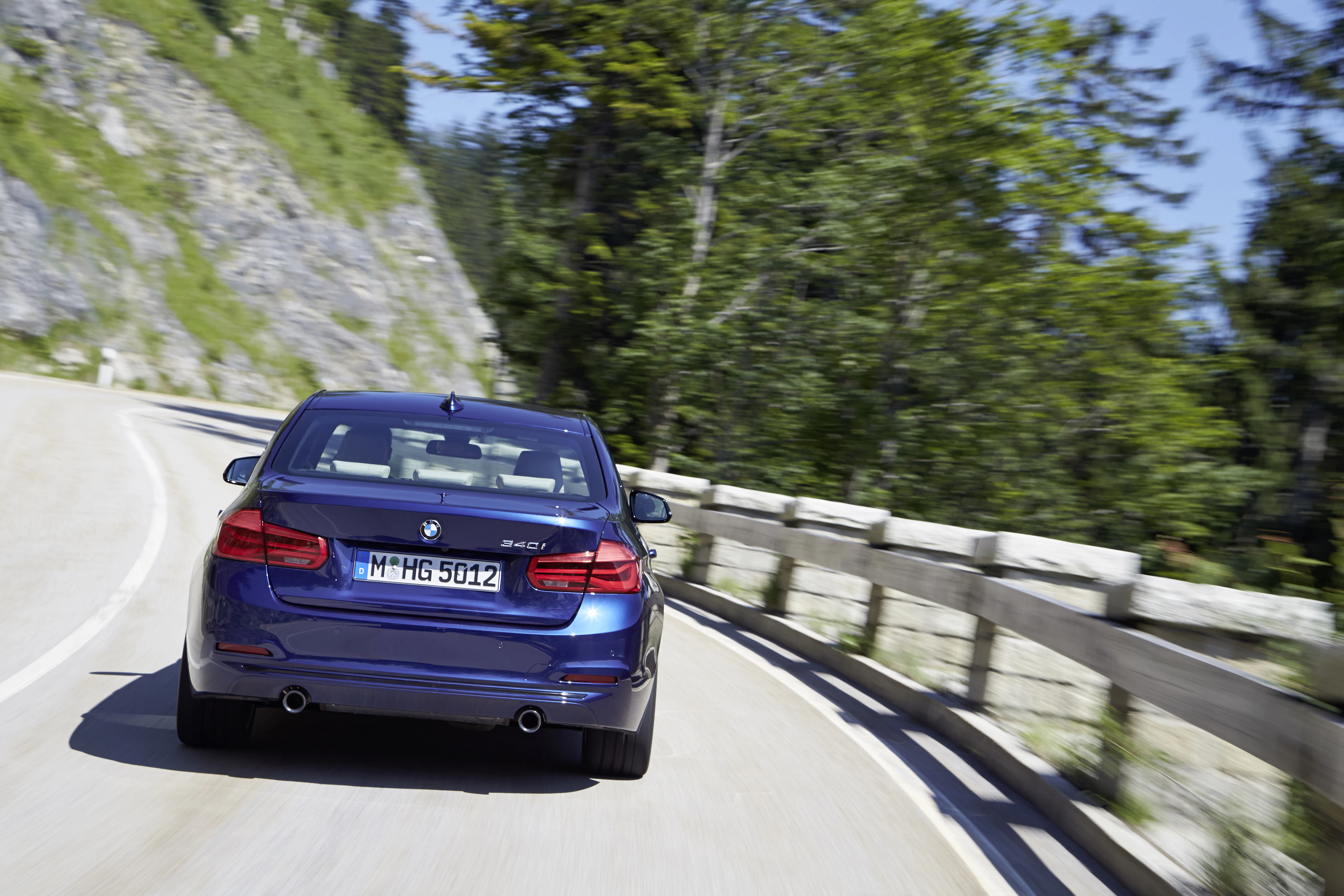 Blaues Auto fährt auf einer kurvigen Bergstraße, umgeben von Bäumen und Felswänden. Lichteinfall erzeugt eine dynamische Szene.