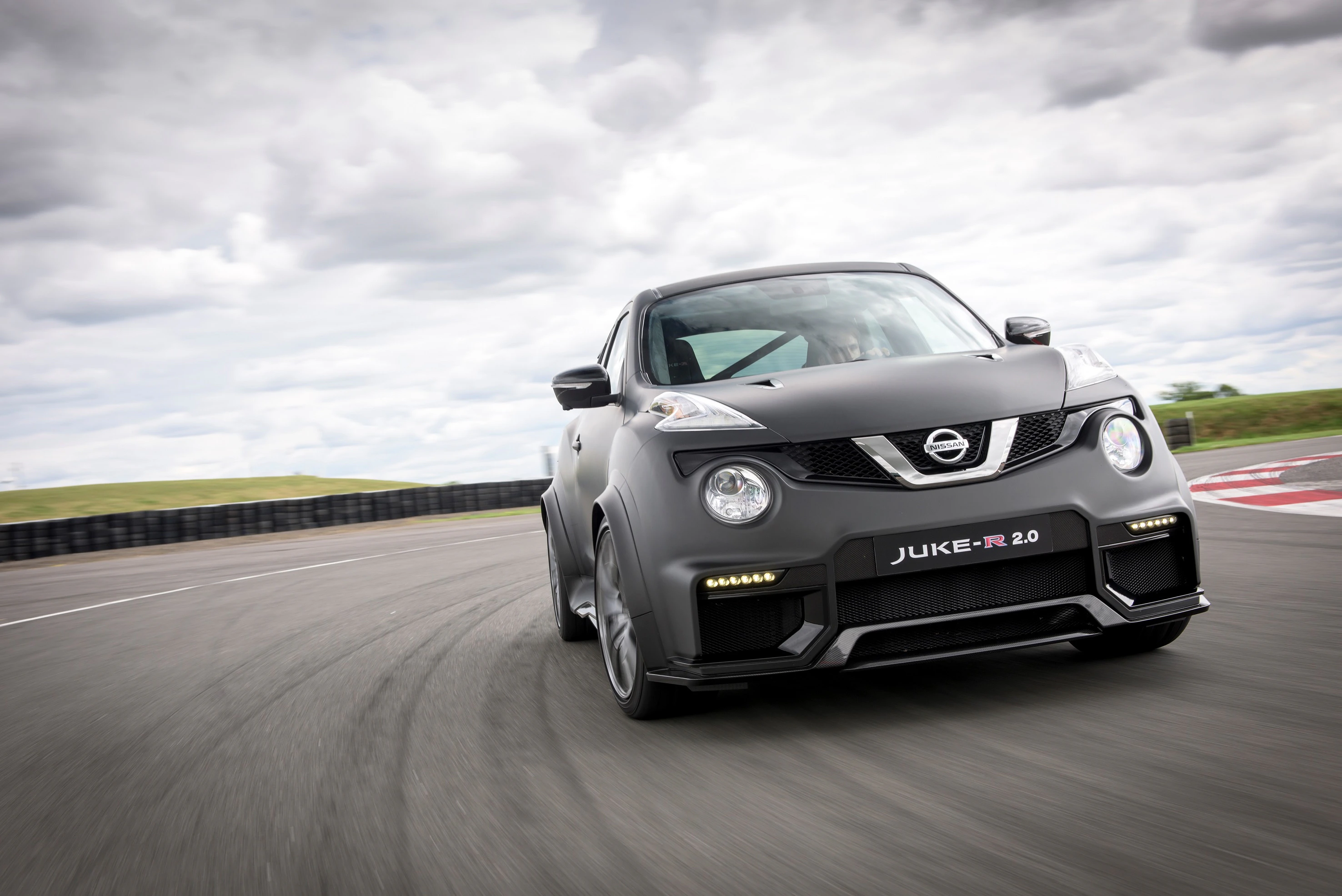 Nissan Juke R 2.0 in mattem Grau fährt dynamisch auf einer Rennstrecke bei bewölktem Himmel. Sportwagen-Design und lebhafte Bewegung.