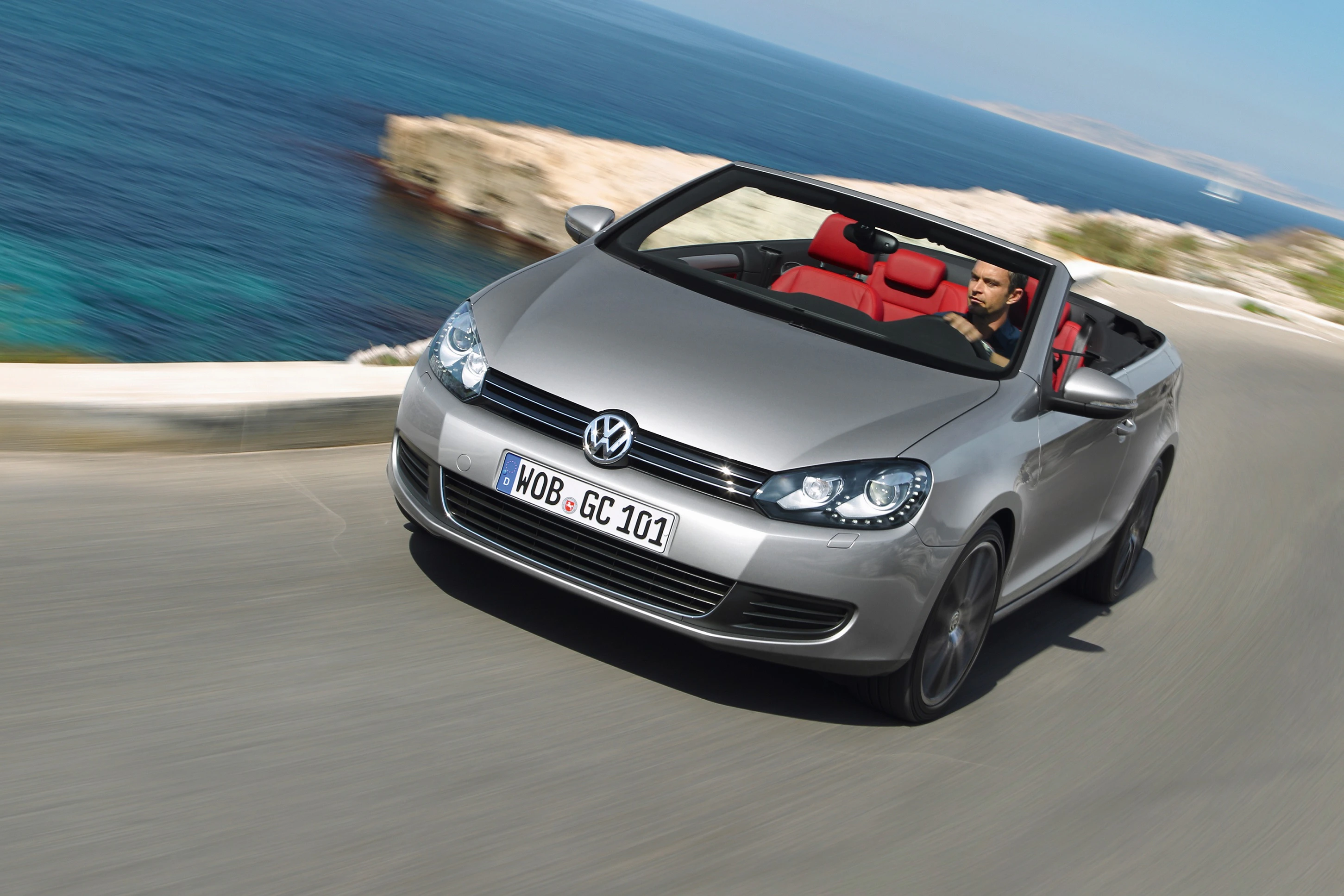 Silbernes Volkswagen Cabrio fährt an einer Küstenstraße entlang, mit einem Fahrer und einem Beifahrer, blaues Meer und Klippen im Hintergrund.