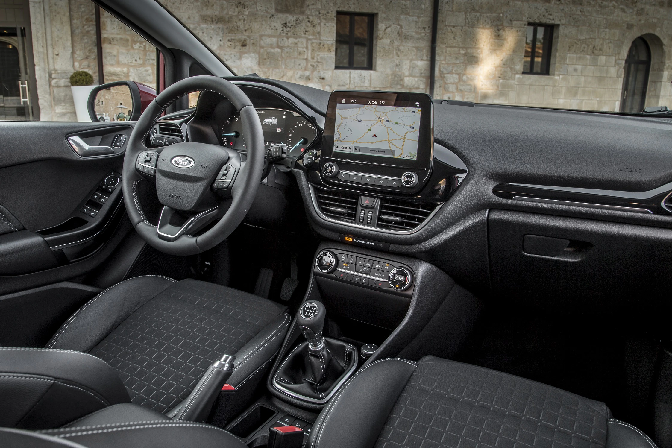 Innenraum eines modernen Autos mit Lenkrad, Touchscreen-Navigation, Schalthebel und schwarzen Sitzen vor einer Steinwandkulisse.