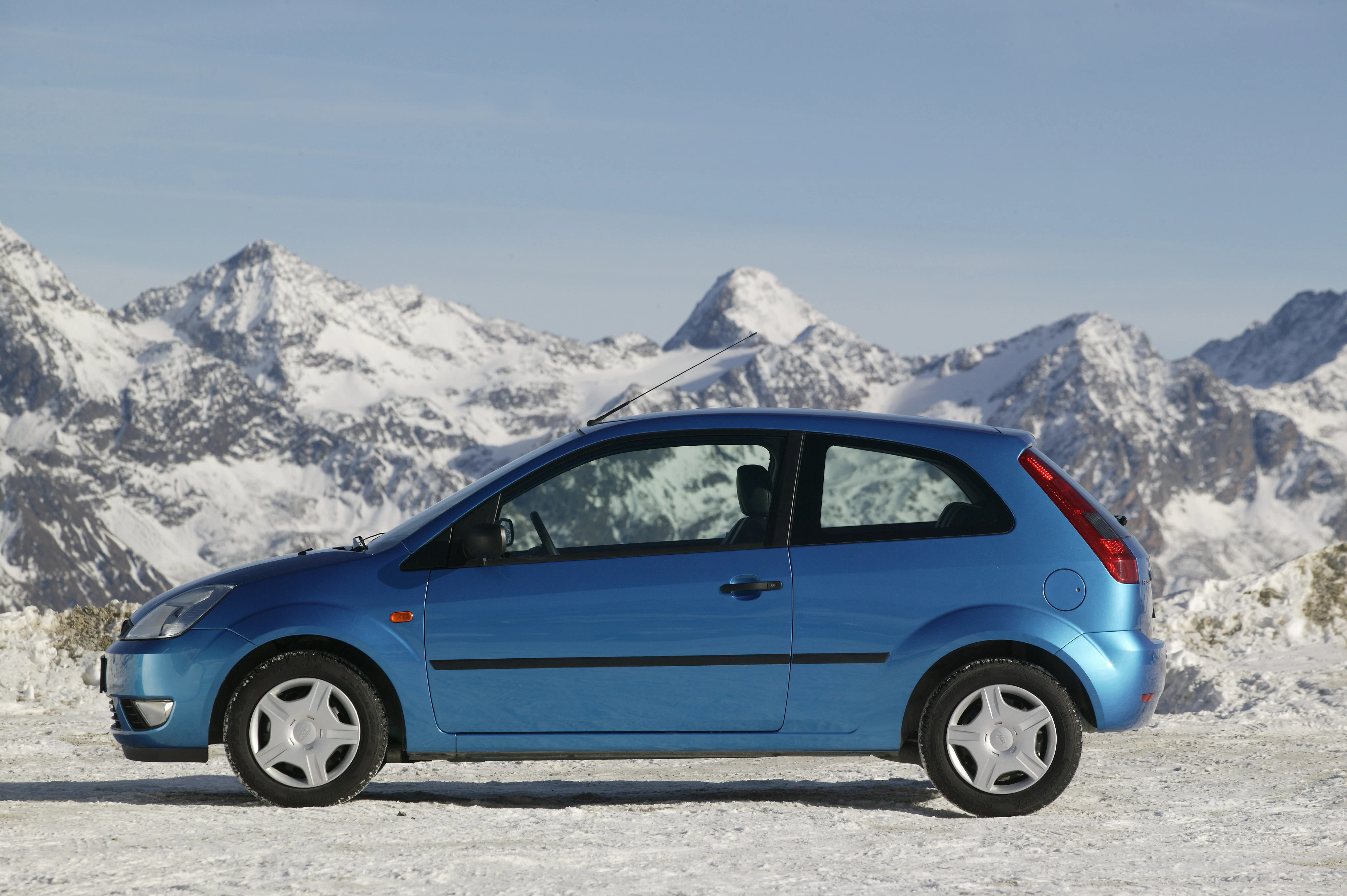 Blaues Auto auf schneebedecktem Boden mit verschneiten Bergen im Hintergrund. Klarer Himmel.