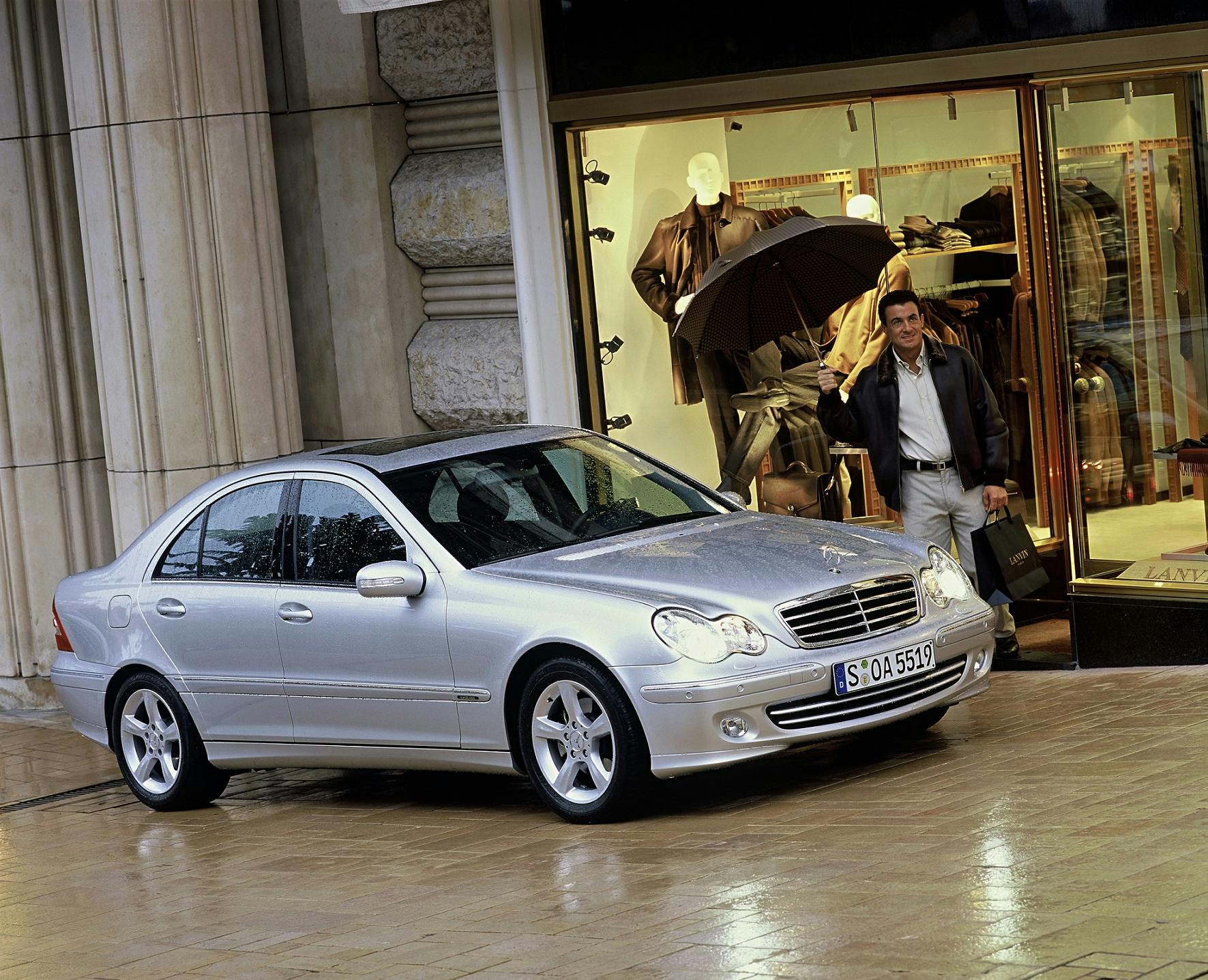 Silberner Mercedes-Benz parkt vor einem eleganten Bekleidungsgeschäft. Ein Mann mit Einkaufstasche und Handy geht aus dem Laden.