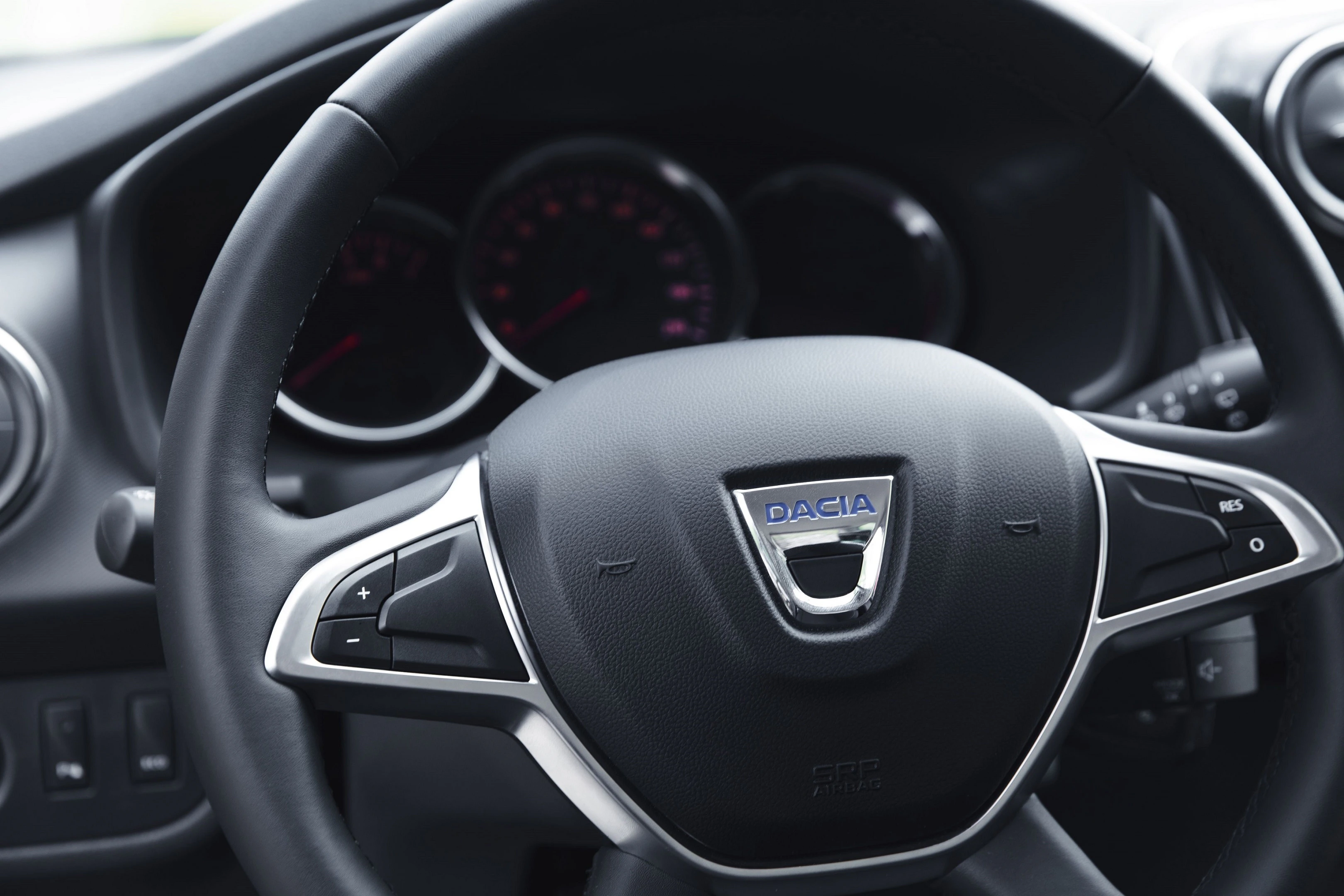 Lenkrad eines Dacia-Fahrzeugs mit Geschwindigkeitsregelungstasten, Tachometer und Armaturenbrettinstrumenten im Hintergrund.