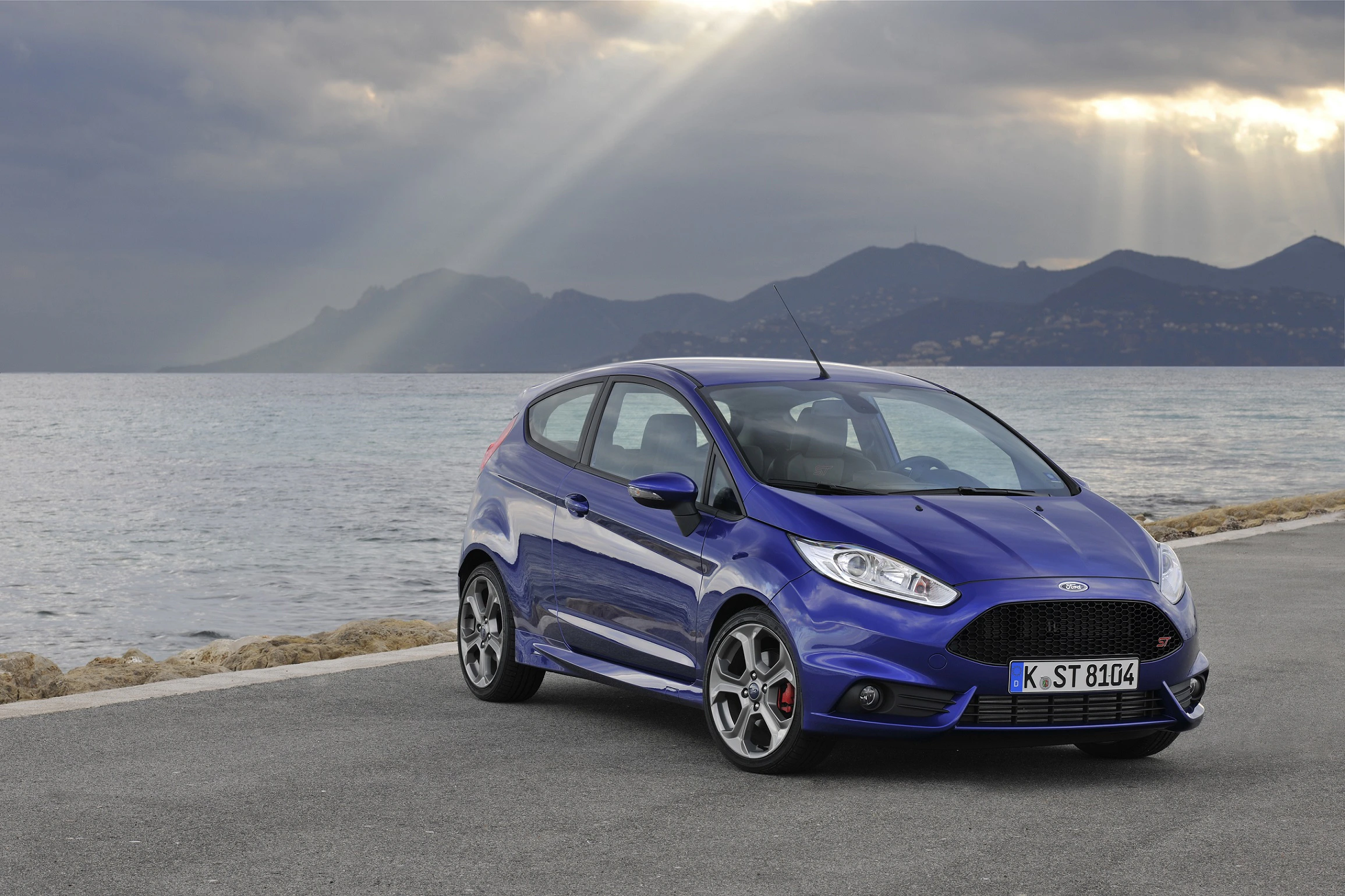 Blauer Ford Fiesta parkt auf Küstenstraße, im Hintergrund Meer und Berge unter bewölktem Himmel mit Sonnenstrahlen.