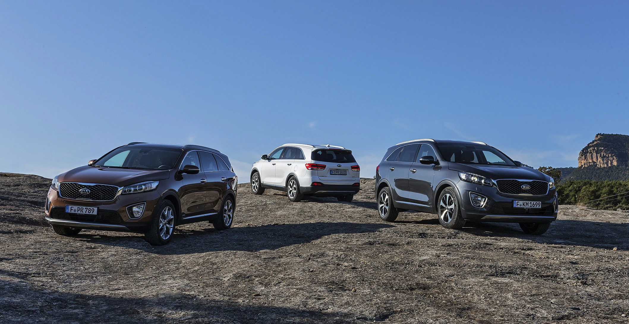 Drei SUVs parken auf felsigem Gelände unter blauem Himmel, ein brauner, ein weißer und ein grauer.