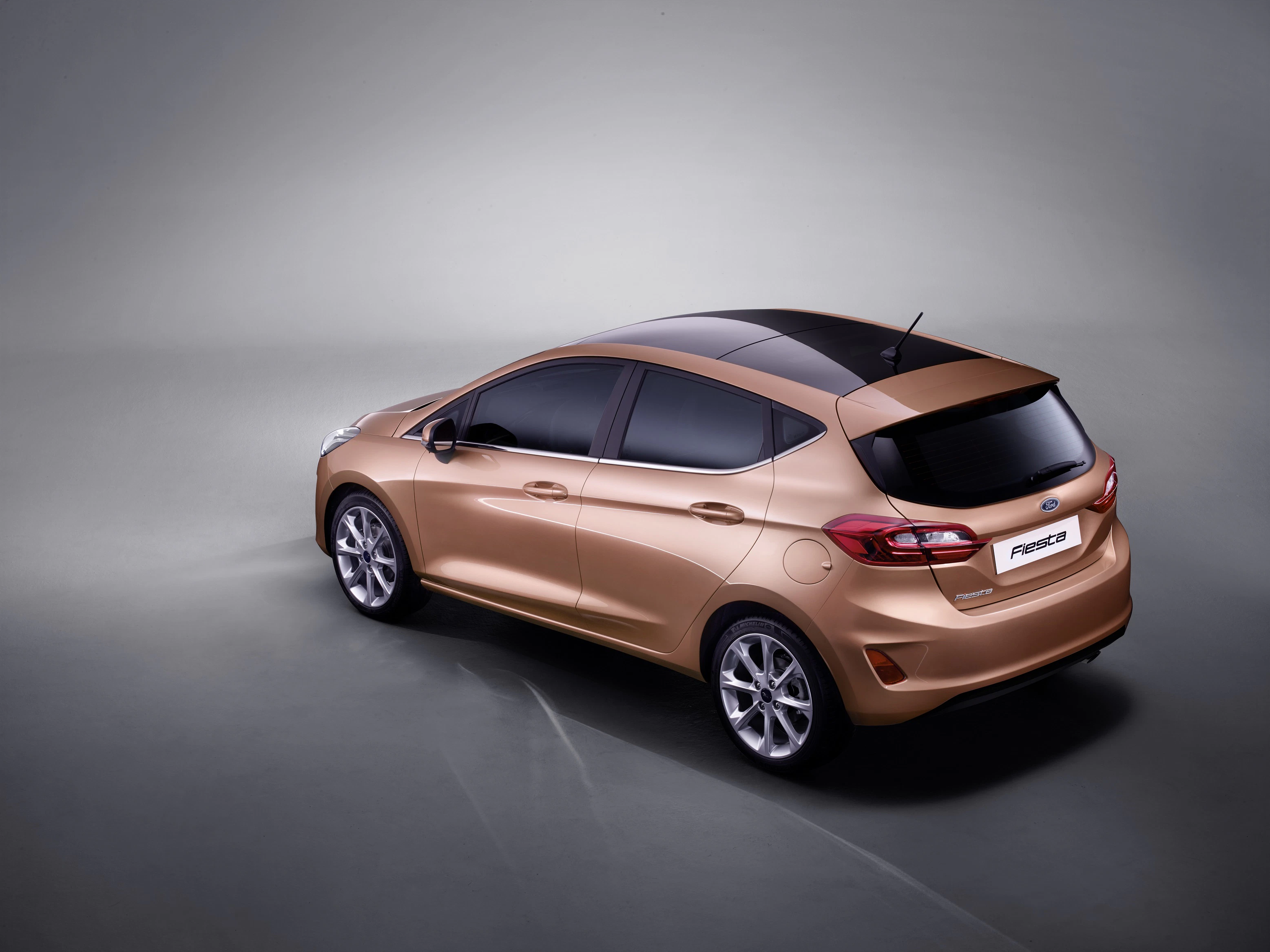 Goldener Kompaktwagen (Ford Fiesta) auf grauem Hintergrund, dreiviertel Heckansicht. Elegantes Design mit getönten Scheiben und Alufelgen.