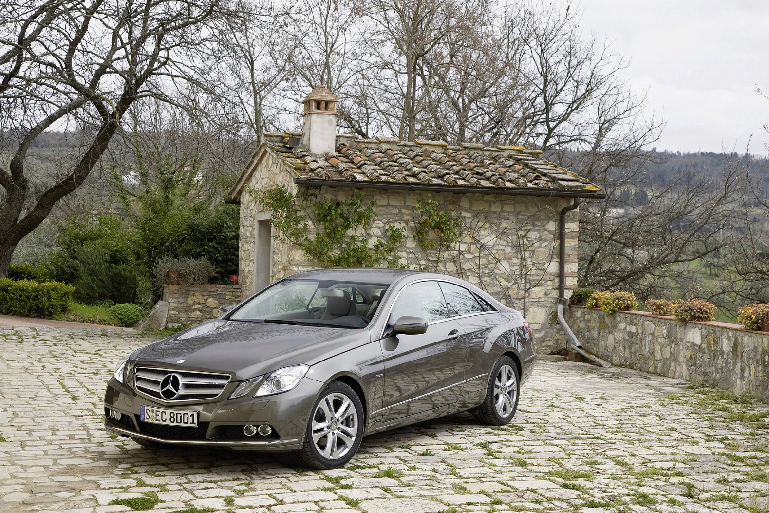 Ein grauer Mercedes-Benz parkt auf einem mit Pflastersteinen bedeckten Hof vor einem Steinhaus mit Ziegeldach.