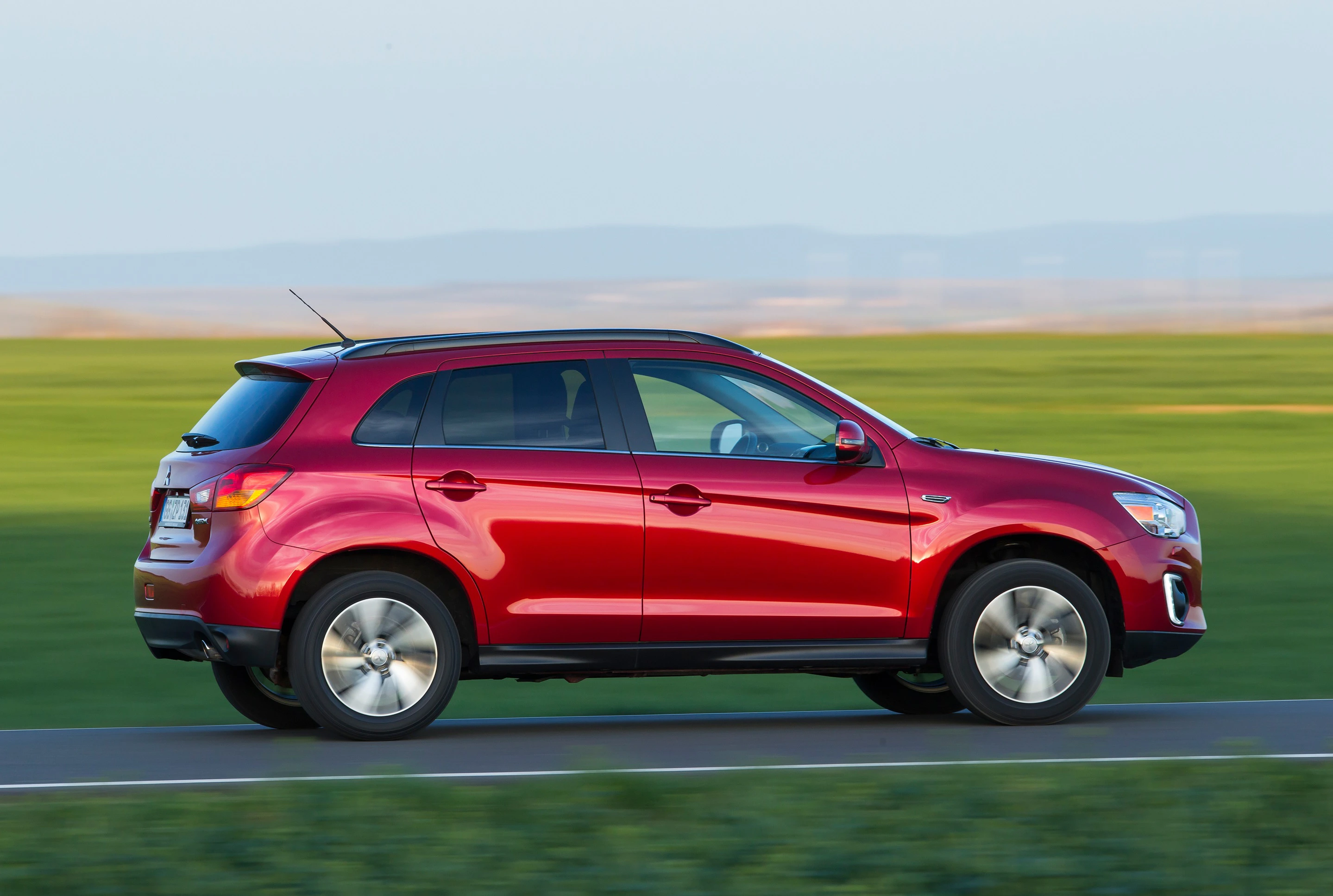 Roter SUV fährt auf einer Landstraße vor grüner Wiesenlandschaft, leichter Horizont im Hintergrund.
