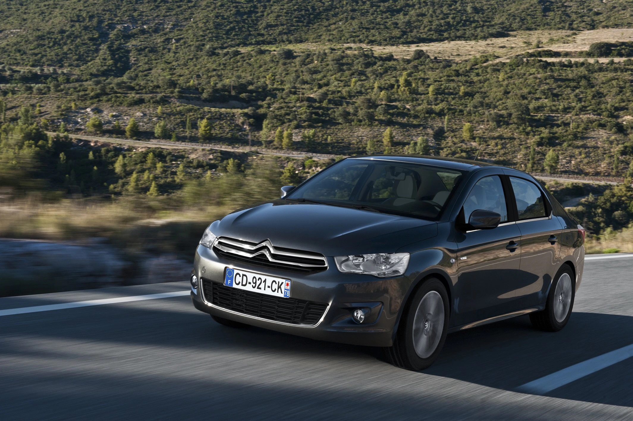 Ein grauer Citroën C4 fährt auf einer Landstraße durch hügelige, grüne Landschaften bei klarem Himmel.