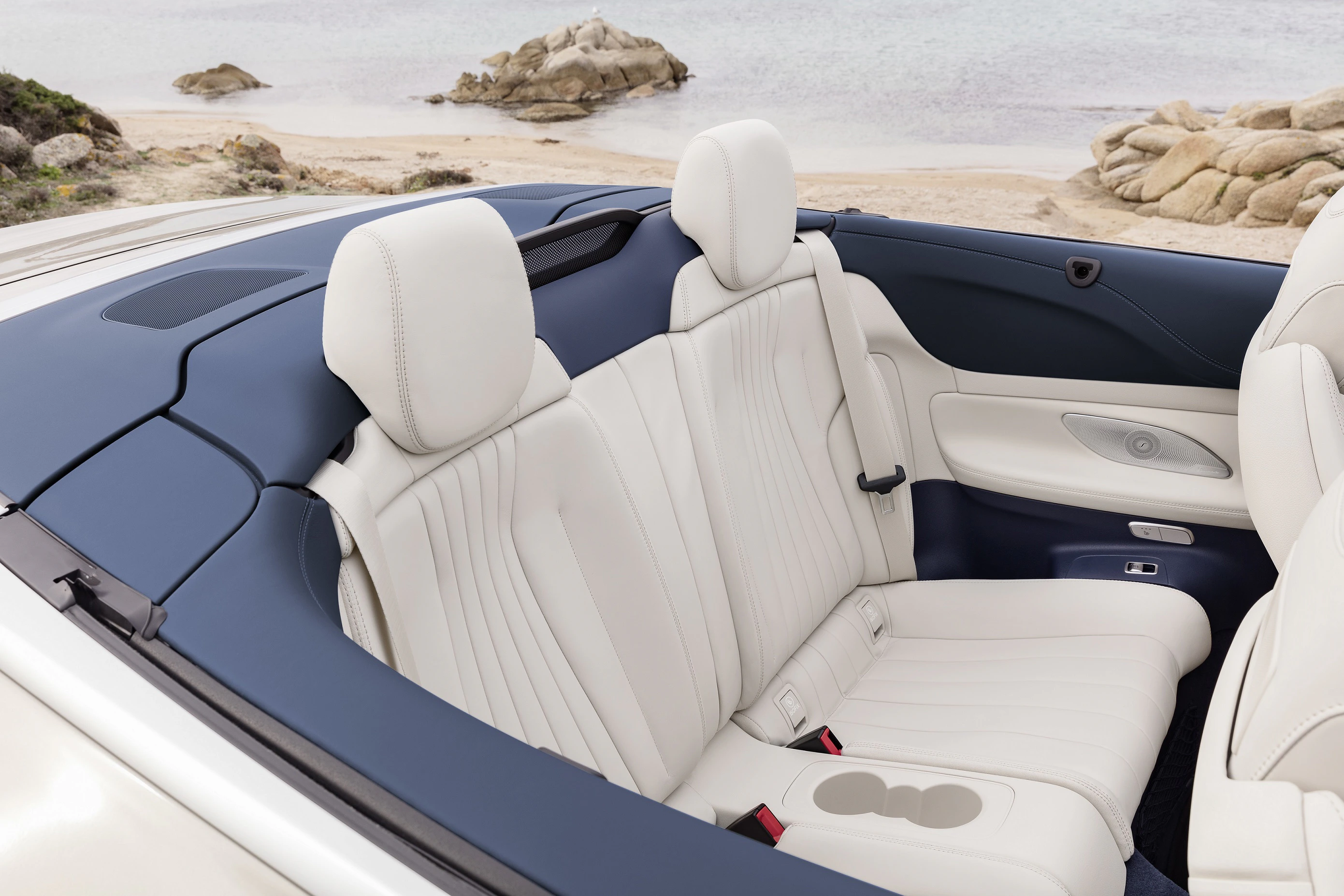 Innenraum eines Cabriolets mit weißen Ledersitzen und blauer Verkleidung. Im Hintergrund ist ein Strand mit Felsen zu sehen.