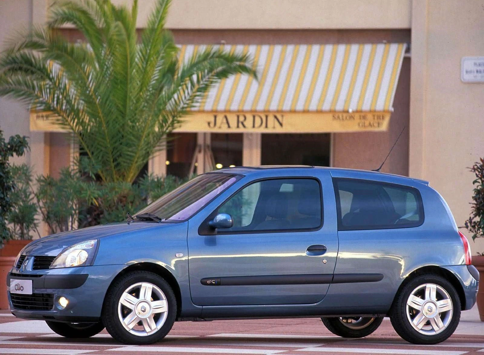 Blauer Kleinwagen steht vor einem Gebäude mit einer Markise "Jardin". Im Hintergrund eine Palme und Pflanzen.