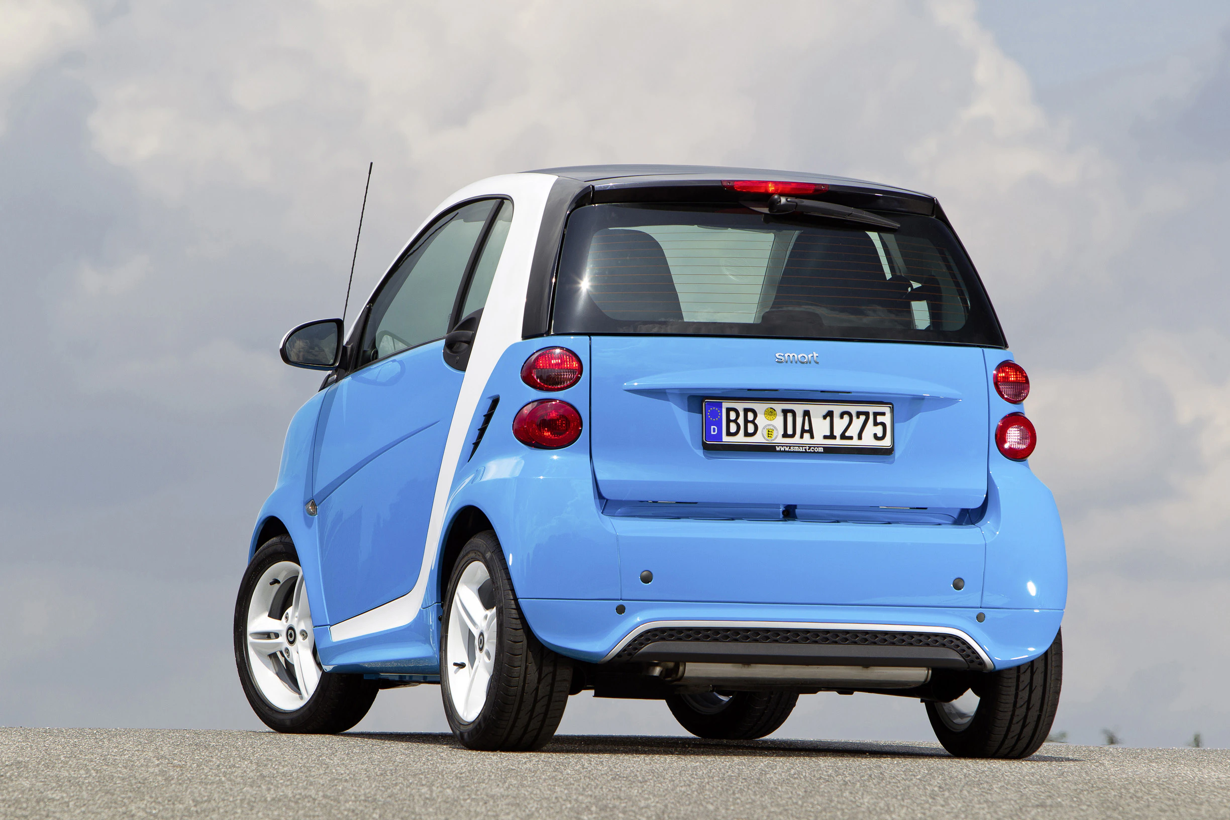 Kleines blaues Smart-Auto von hinten auf einer Straße, Nummernschild sichtbar, bedeckter Himmel im Hintergrund.