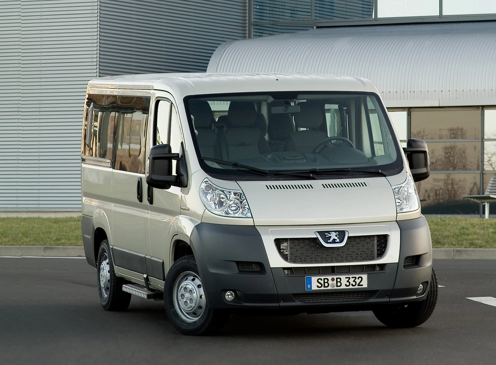 Silberner Peugeot Kleinbus, parkt vor einem modernen Gebäude mit Glas- und Stahlfassade. Lizenznummer sichtbar: SB:B 332.