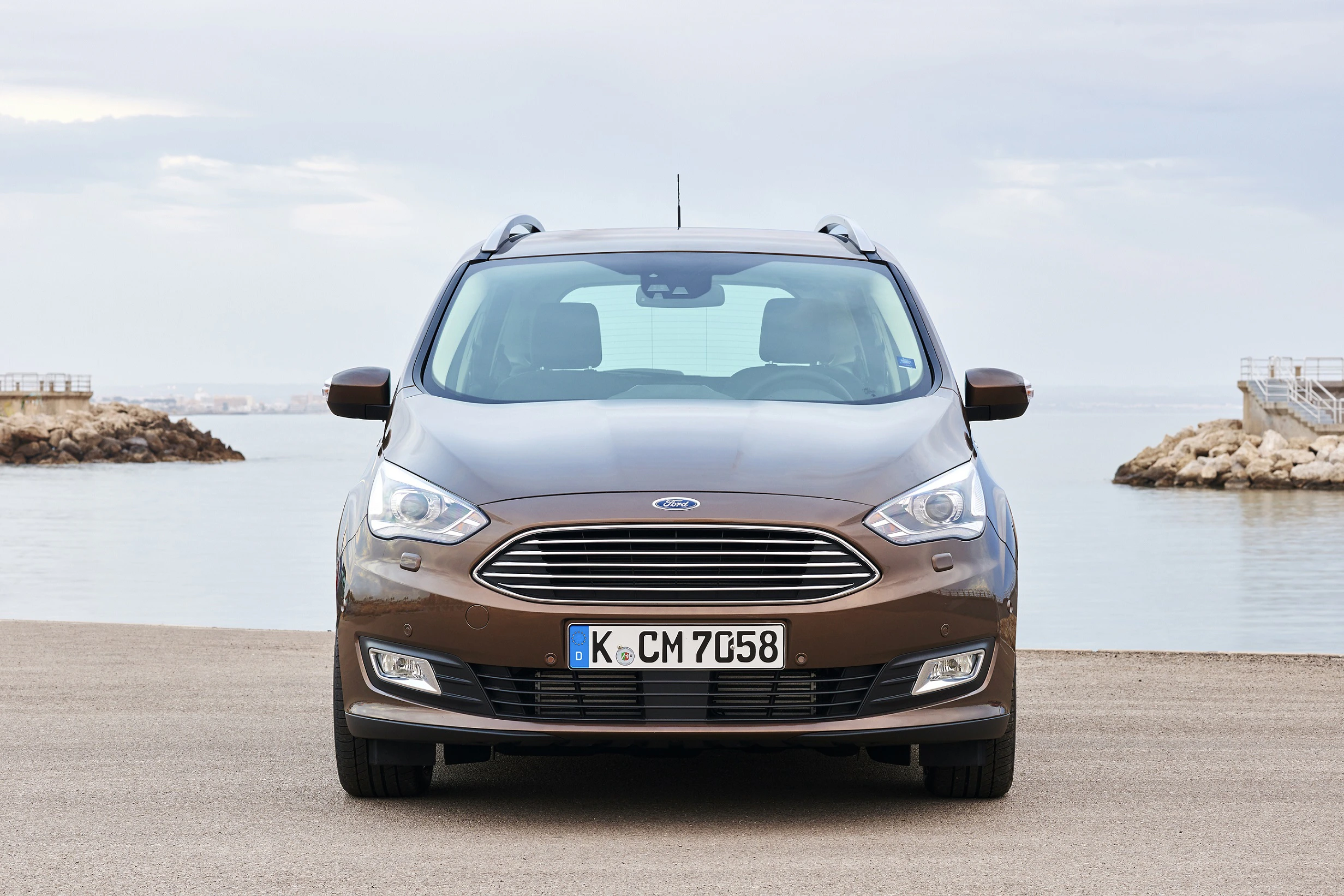 Brauner Ford C-Max, von vorne, geparkt vor einem Hafen mit klarem Himmel und ruhigem Wasser im Hintergrund.