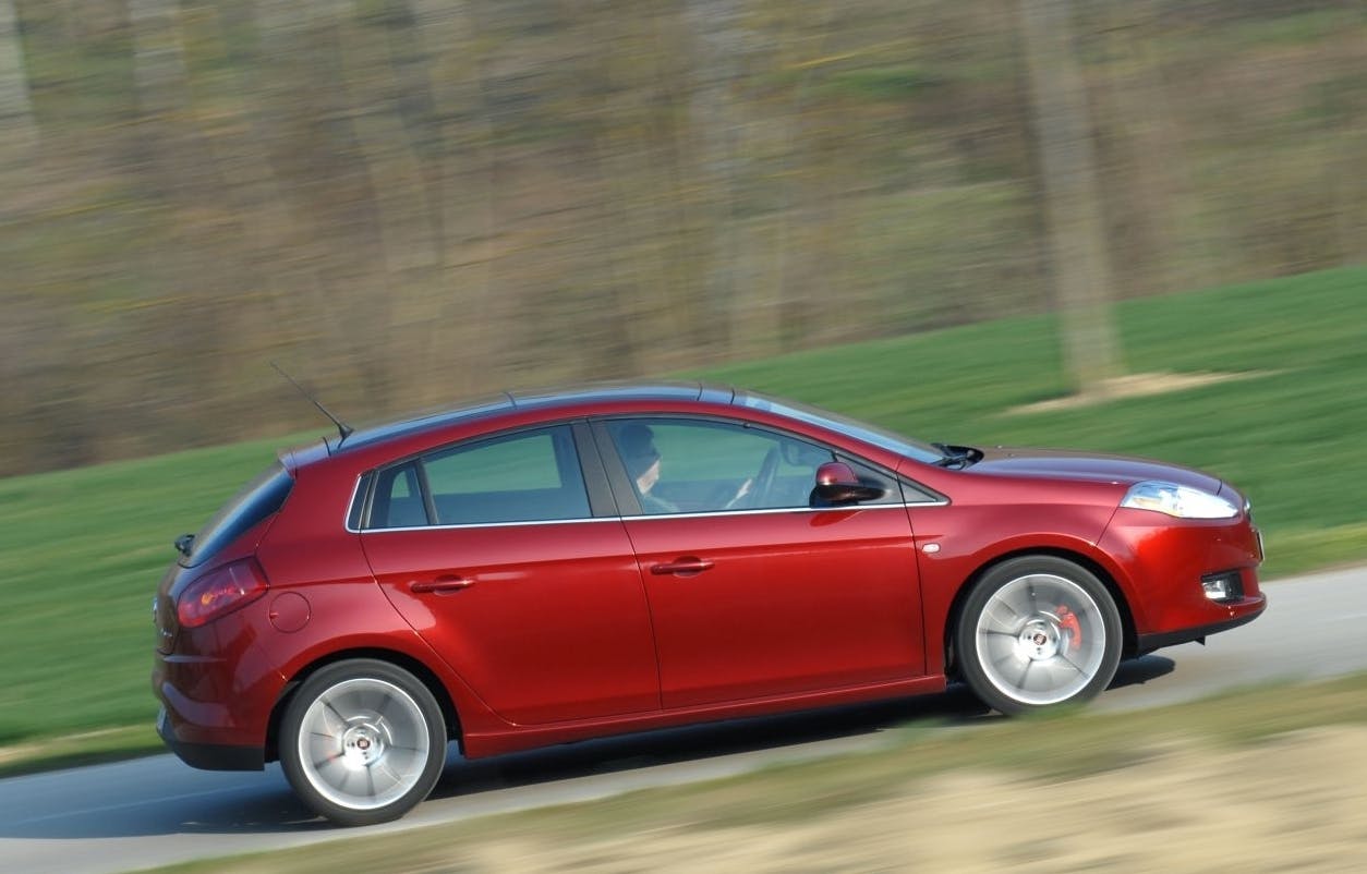 Roter Kleinwagen fährt schnell auf einer Straße, umgeben von grüner Landschaft und unscharfem Hintergrund.
