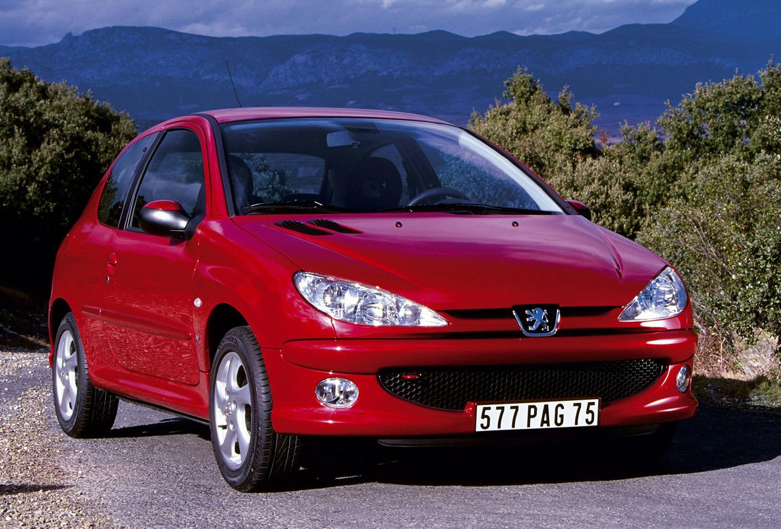 Roter Peugeot 206 steht auf einer Straße vor einer Berglandschaft. Das Auto zeigt französische Nummernschilder und hat ein sportliches Design.