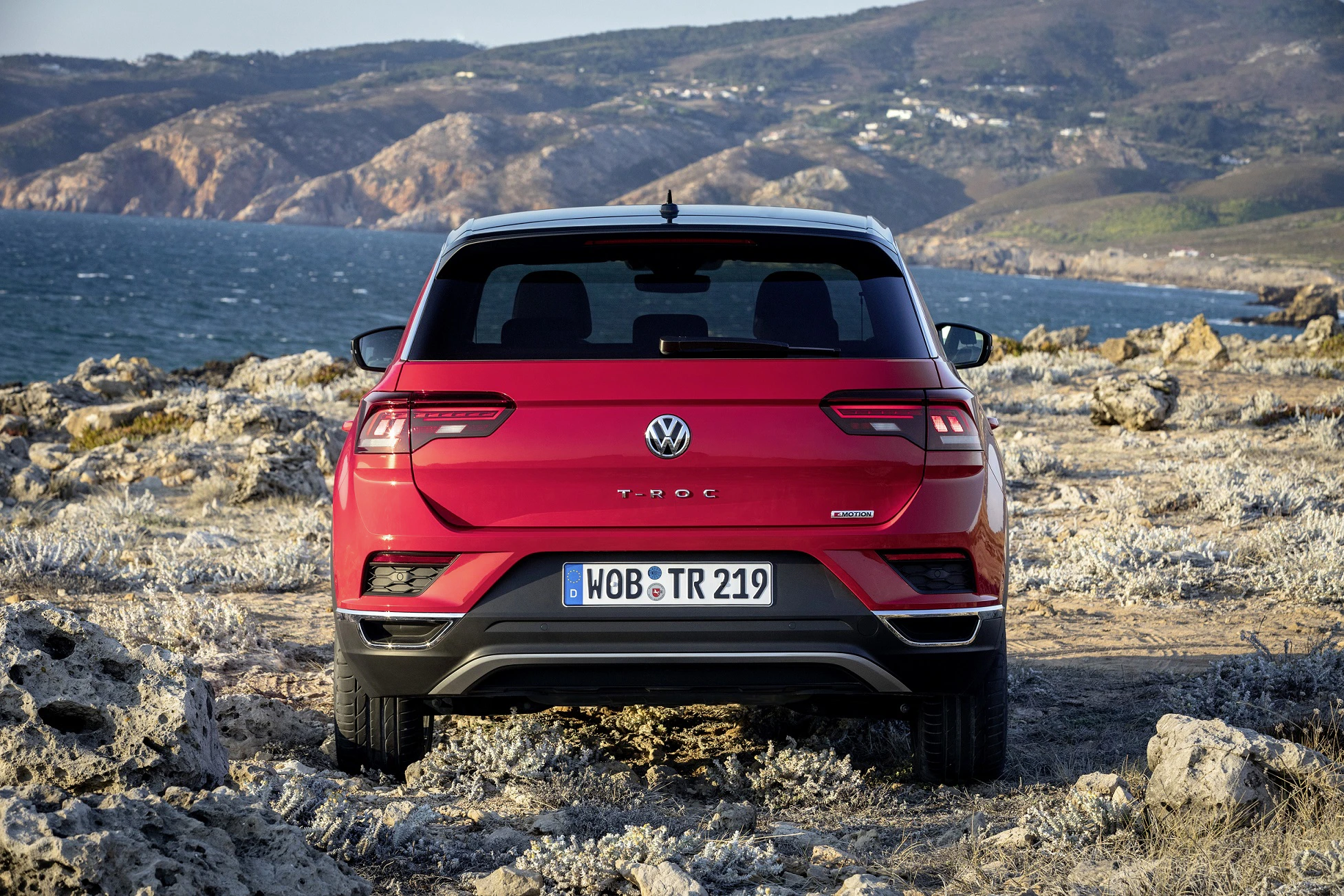 Roter Volkswagen T-Roc mit Meeresblick, geparkt auf felsigem Gelände, im Hintergrund Hügel und Küste unter blauem Himmel.