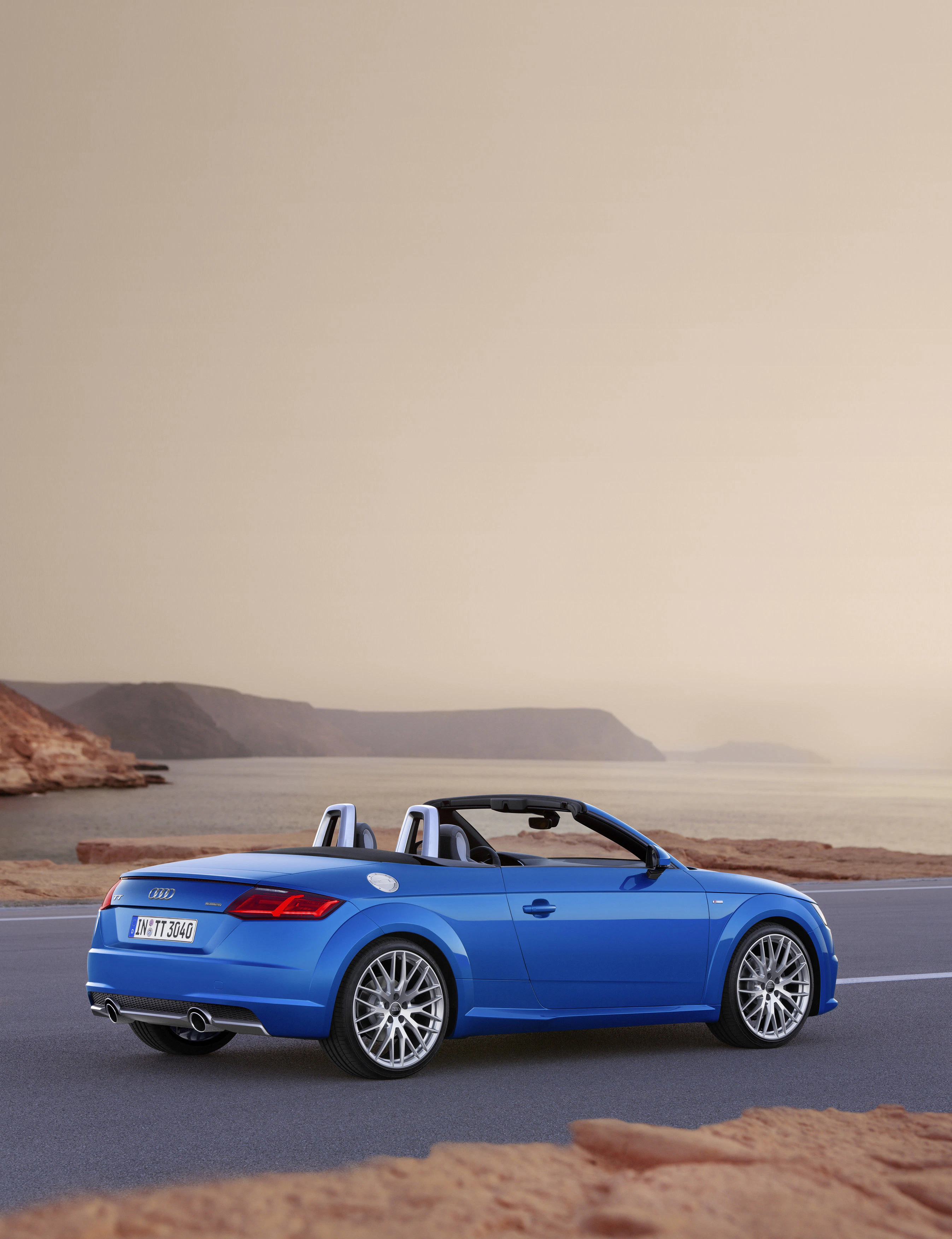 Blauer Audi TT Cabriolet parkt auf einer Küstenstraße bei Sonnenuntergang. Hintergrund mit Felsen und Meer.