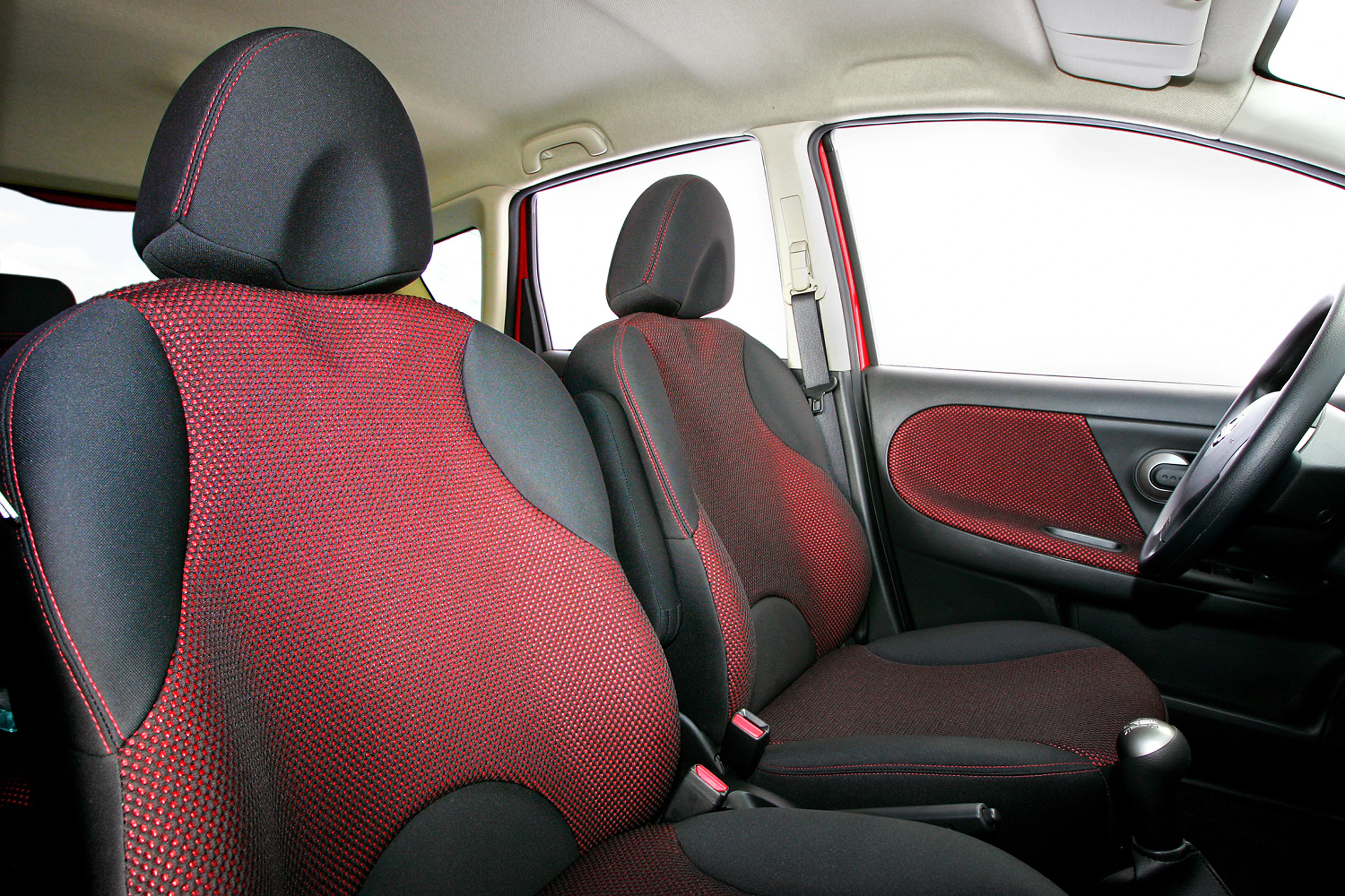 Innenraum eines Autos mit zwei Vordersitzen in schwarzem und rotem Stoff. Helles Cockpit mit sichtbaren Türen und Fenster.