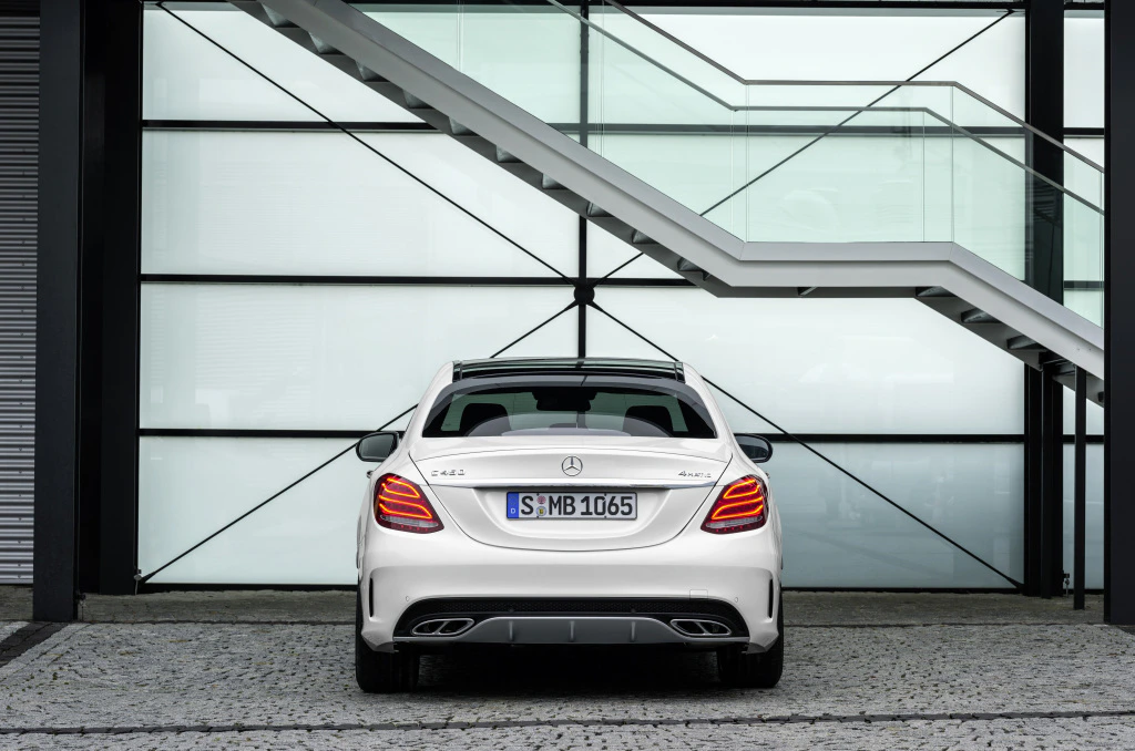 Ein weißer Mercedes-Benz C-Klasse AMG steht vor einer modernen Glasfassade, hinter einem diagonalen Treppengeländer geparkt.