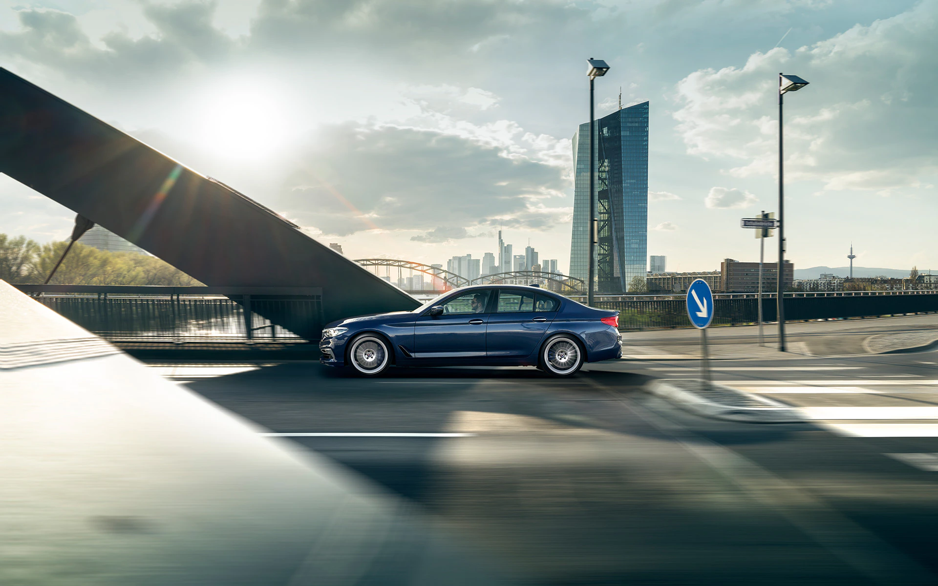 Blaues Auto auf Brücke, mit moderner Stadtlandschaft im Hintergrund bei Sonnenuntergang, Wolken und Wolkenkratzer sichtbar.