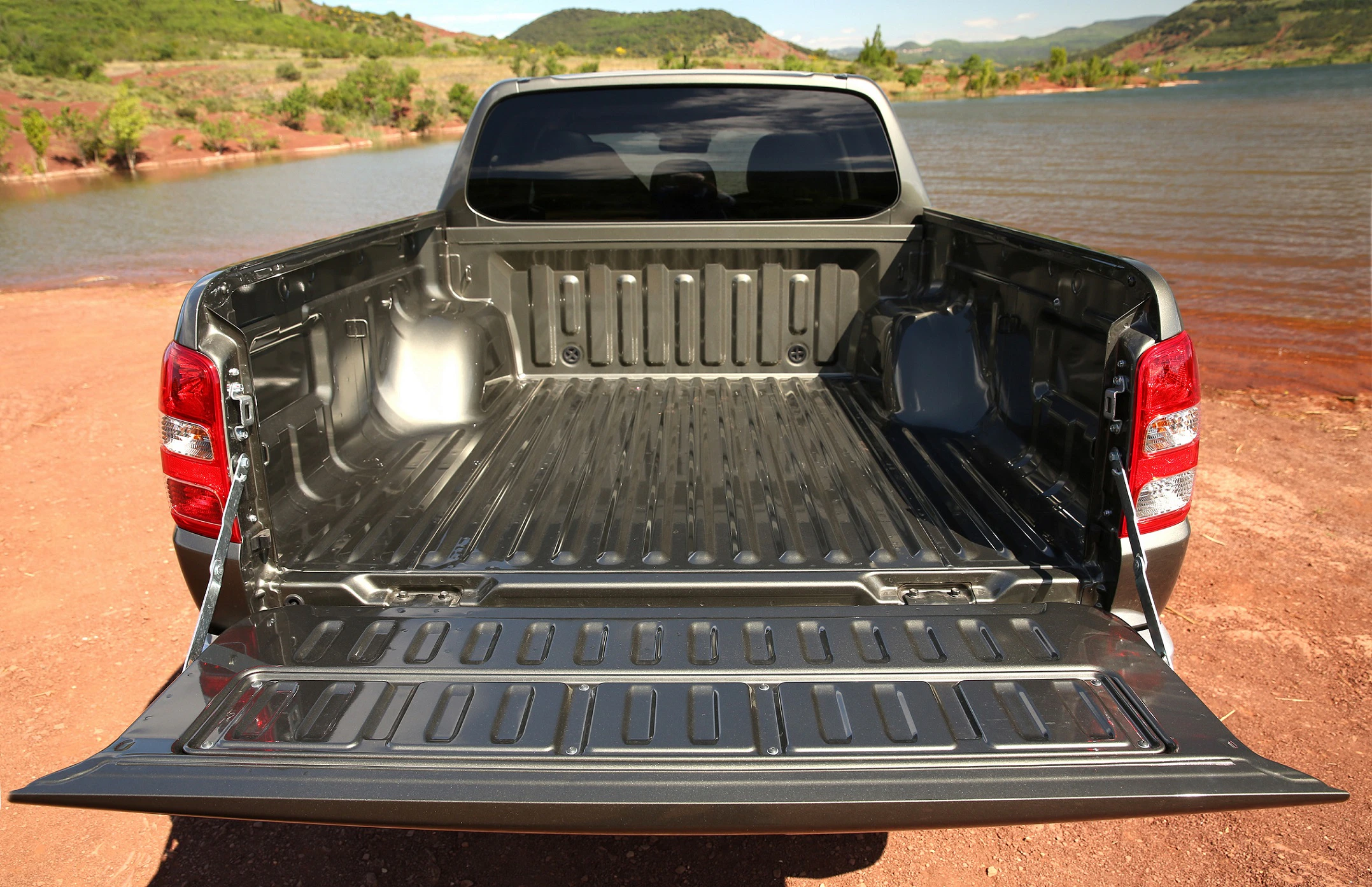 Leerer Laderaum eines silbernen Pickup-Trucks, geöffnet, mit Blick auf einen See und grüne Hügel im Hintergrund.