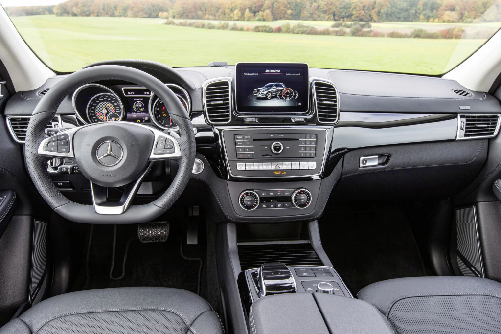 Innenraum eines Mercedes-SUVs mit Lenkrad, Tachometer, Infotainment-Bildschirm, Mittelkonsole und schwarzen Ledersitzen, Blick nach draußen ins Grüne.