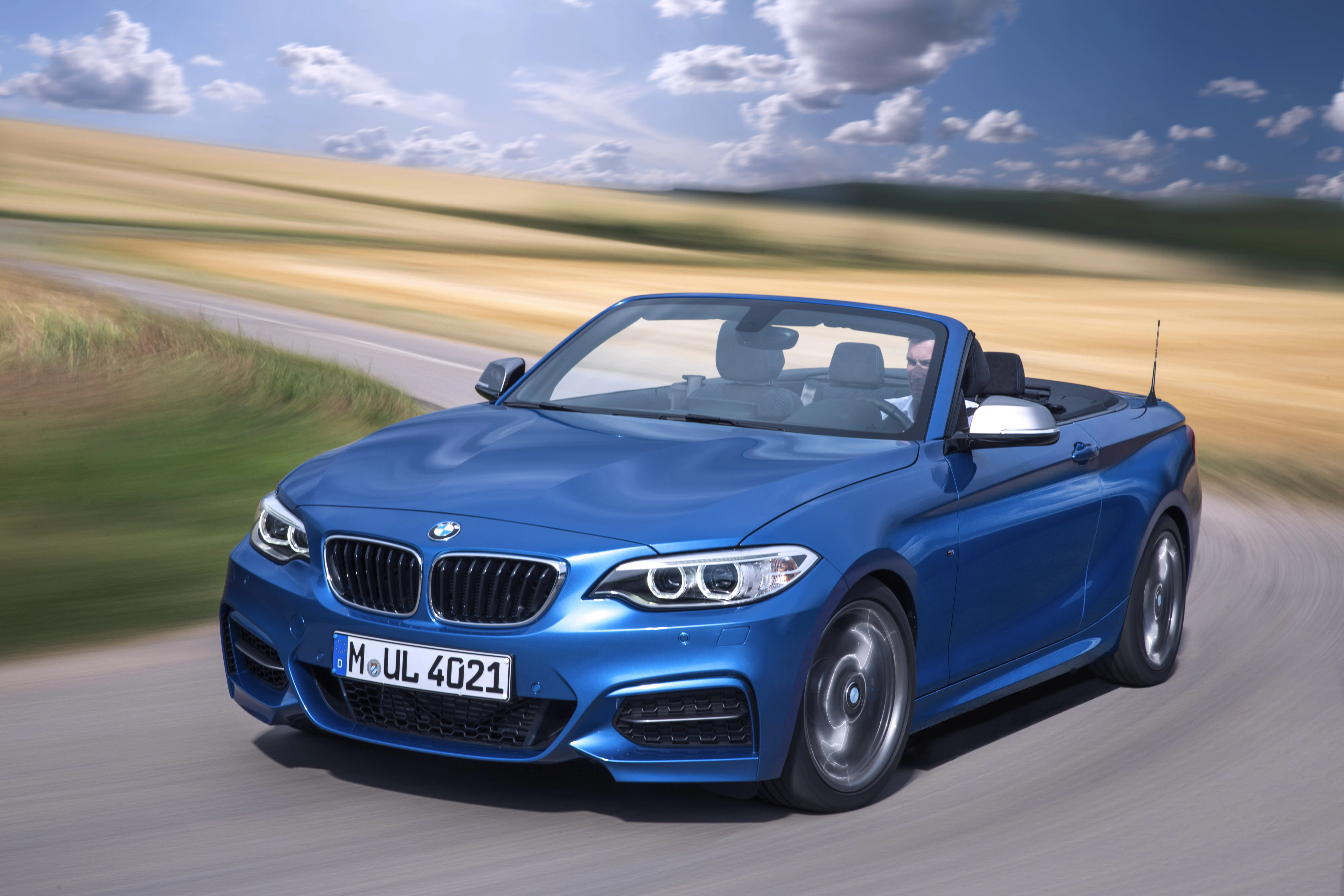Blaues Cabrio fährt auf einer kurvigen Landstraße durch eine ländliche Landschaft bei sonnigem Wetter.