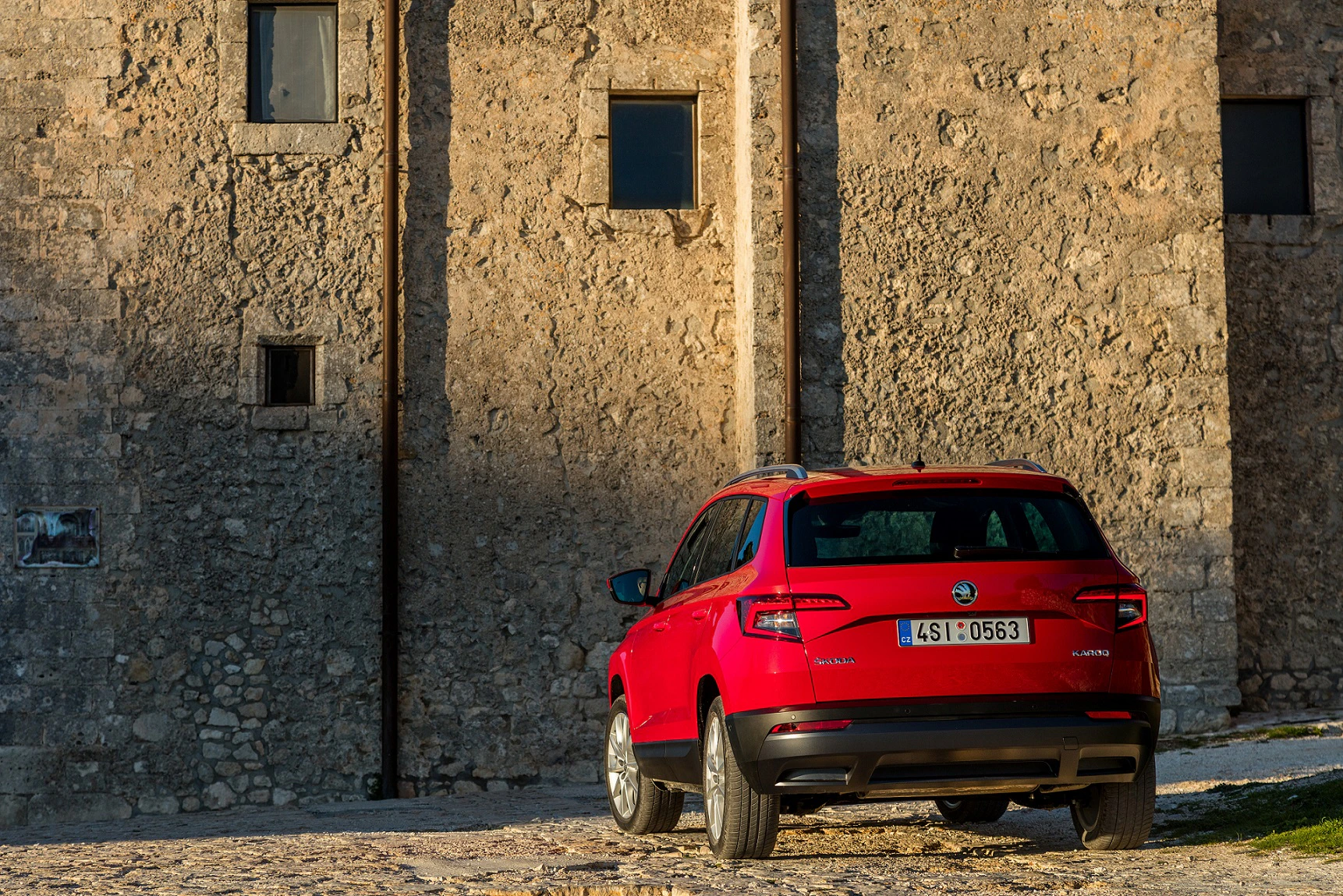 Roter SUV parkt vor einer alten Steinmauer mit kleinen Fenstern im Sonnenlicht.
