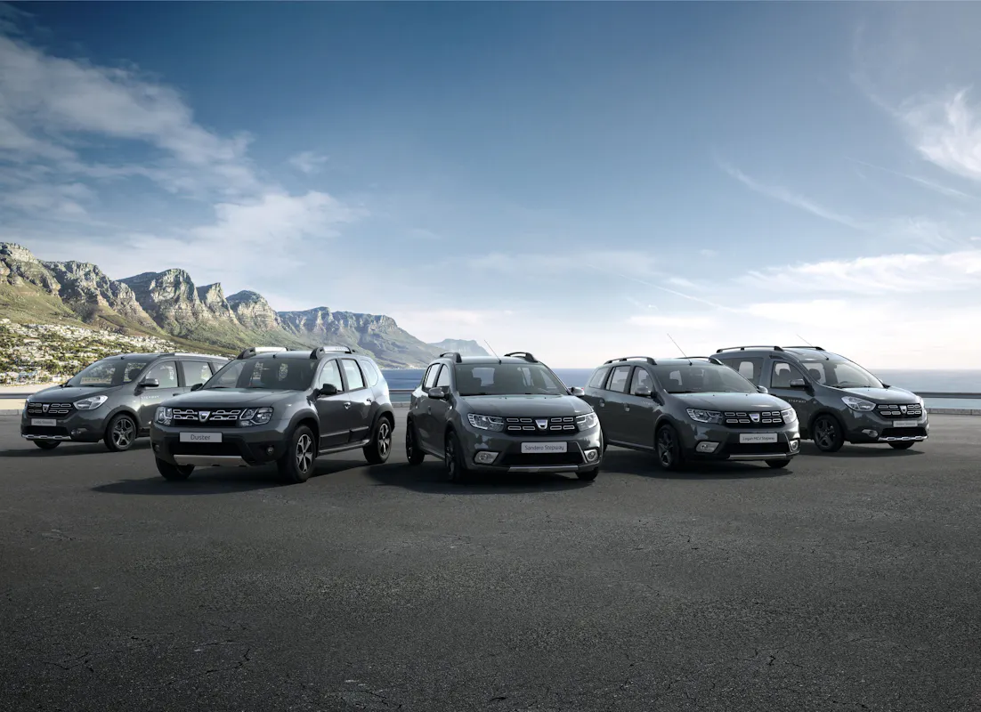 Sechs graue SUVs auf einer Küstenstraße mit Bergen im Hintergrund unter klarem Himmel.