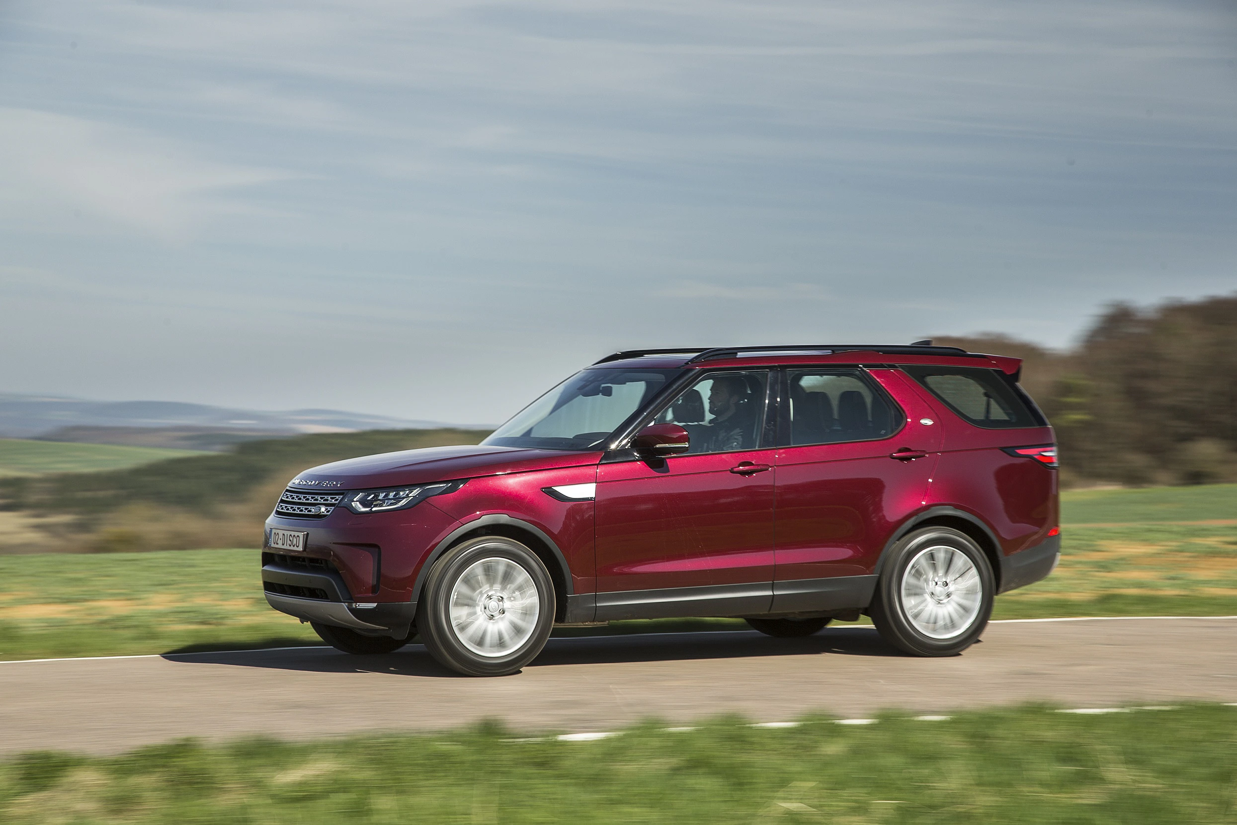 Ein roter SUV fährt auf einer Landstraße durch eine grüne Landschaft bei bewölktem Himmel.