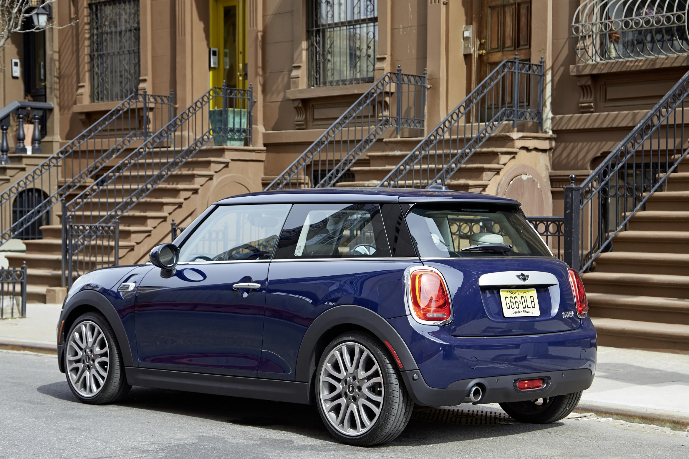 Blauer Mini Cooper parkt vor einem Wohngebäude mit Treppen und Schmiedeeisengeländern auf einer städtischen Straße.