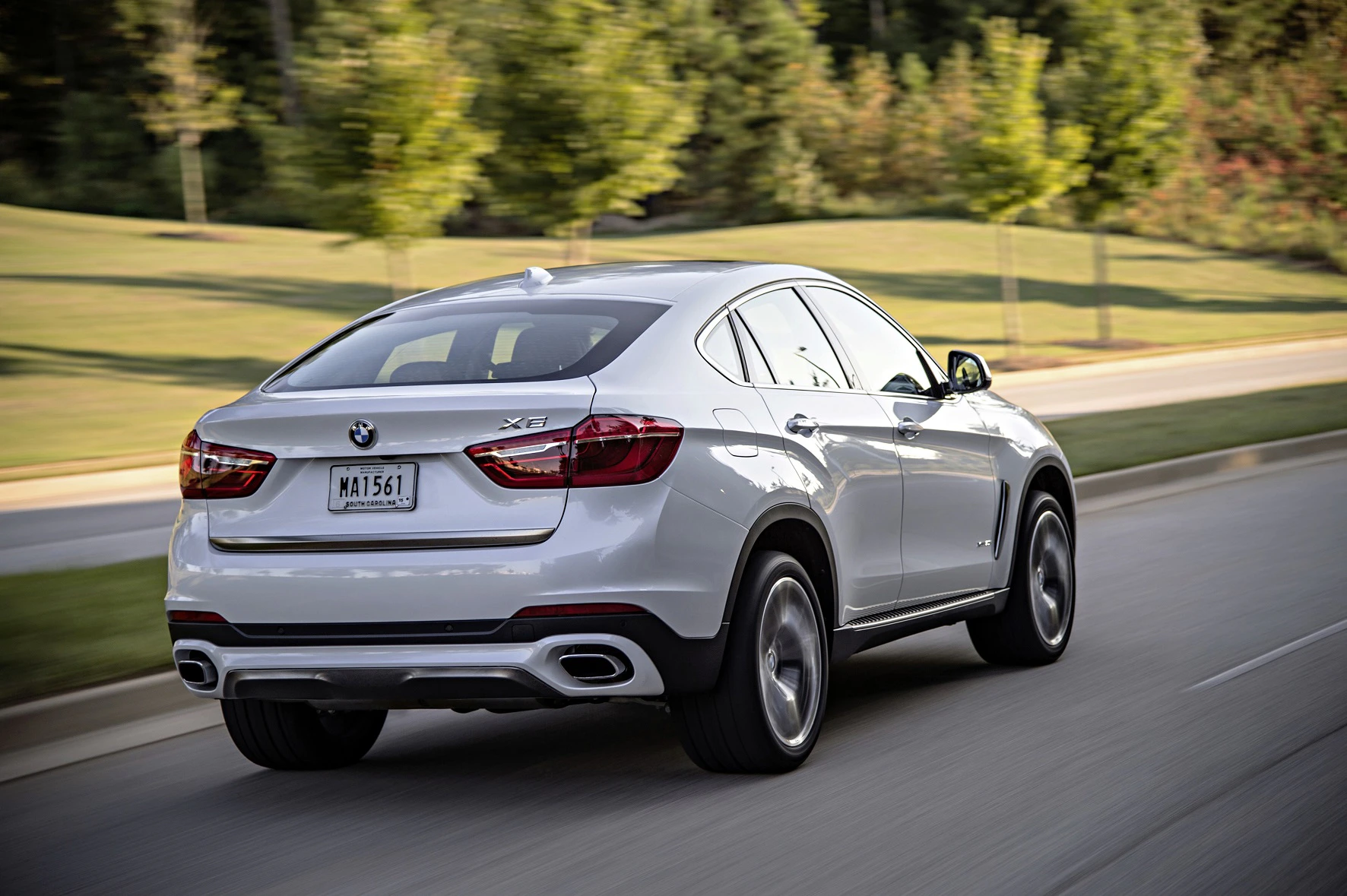 Silberner BMW X6 fährt auf einer Straße in grüner Umgebung. Rückansicht des Autos, Bäume im Hintergrund.