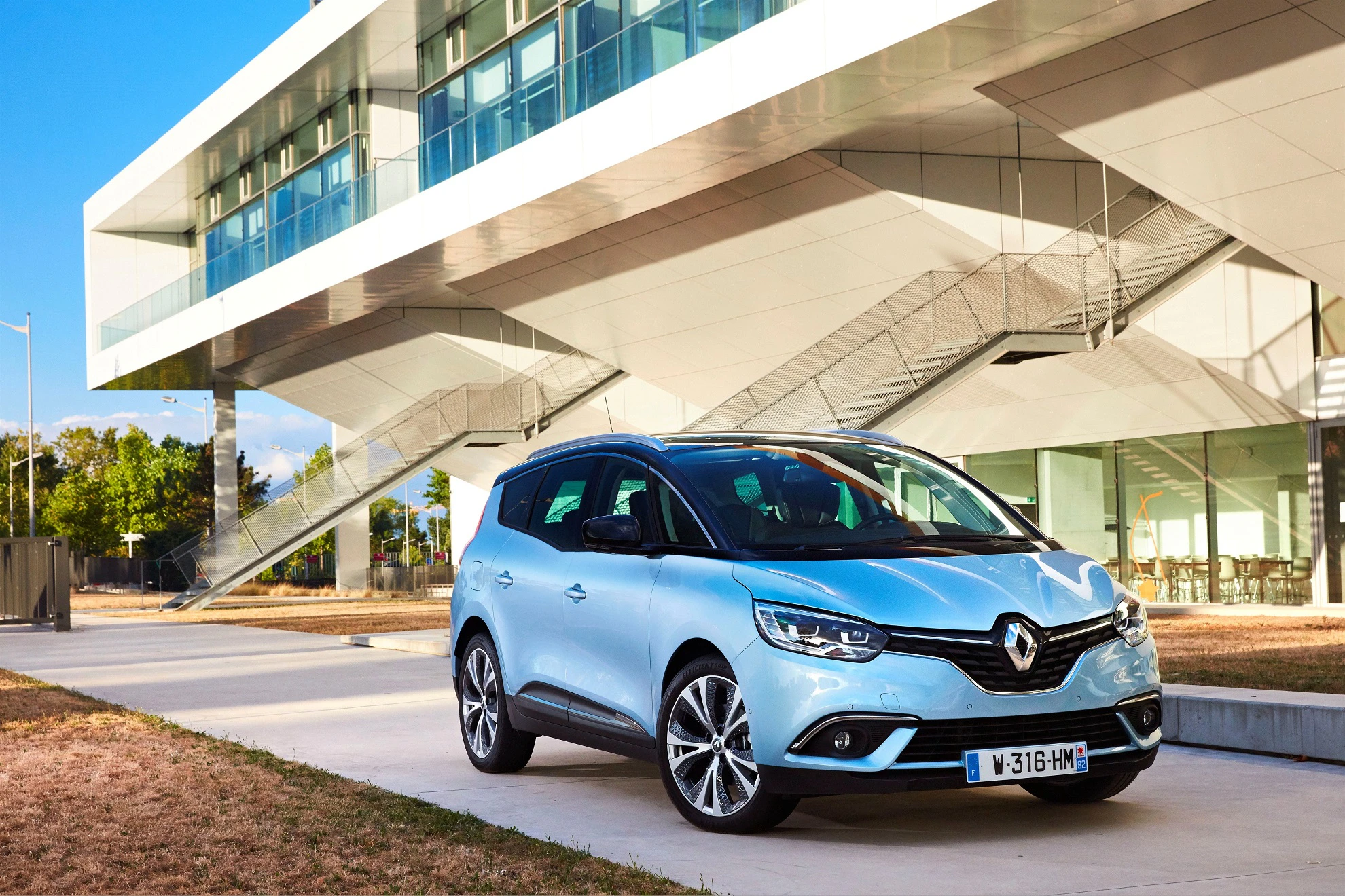 Ein silberner Renault Minivan steht vor einem modernen, weißen Gebäude mit Glasfassaden und schrägen Treppen.