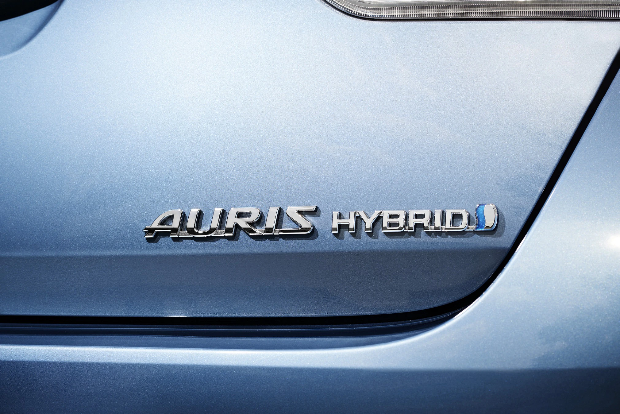 Nahaufnahme eines blauen Autos mit dem Emblem "AURIS HYBRID" auf der Heckklappe, das auf den Hybridantrieb hinweist.