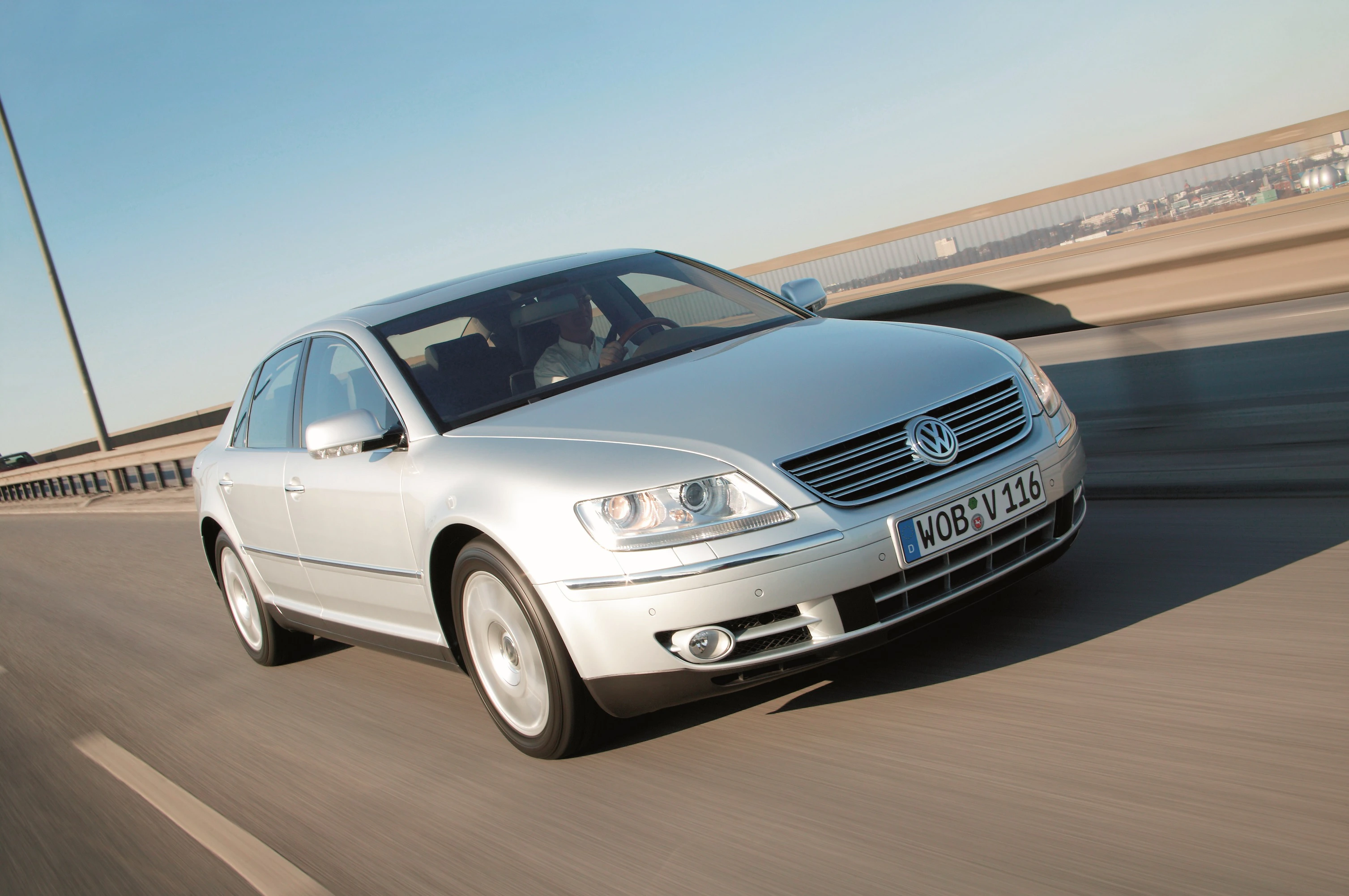 Silberner VW Phaeton fährt auf einer Autobahnbrücke bei Sonnenschein. Vorderansicht, leicht seitlich.