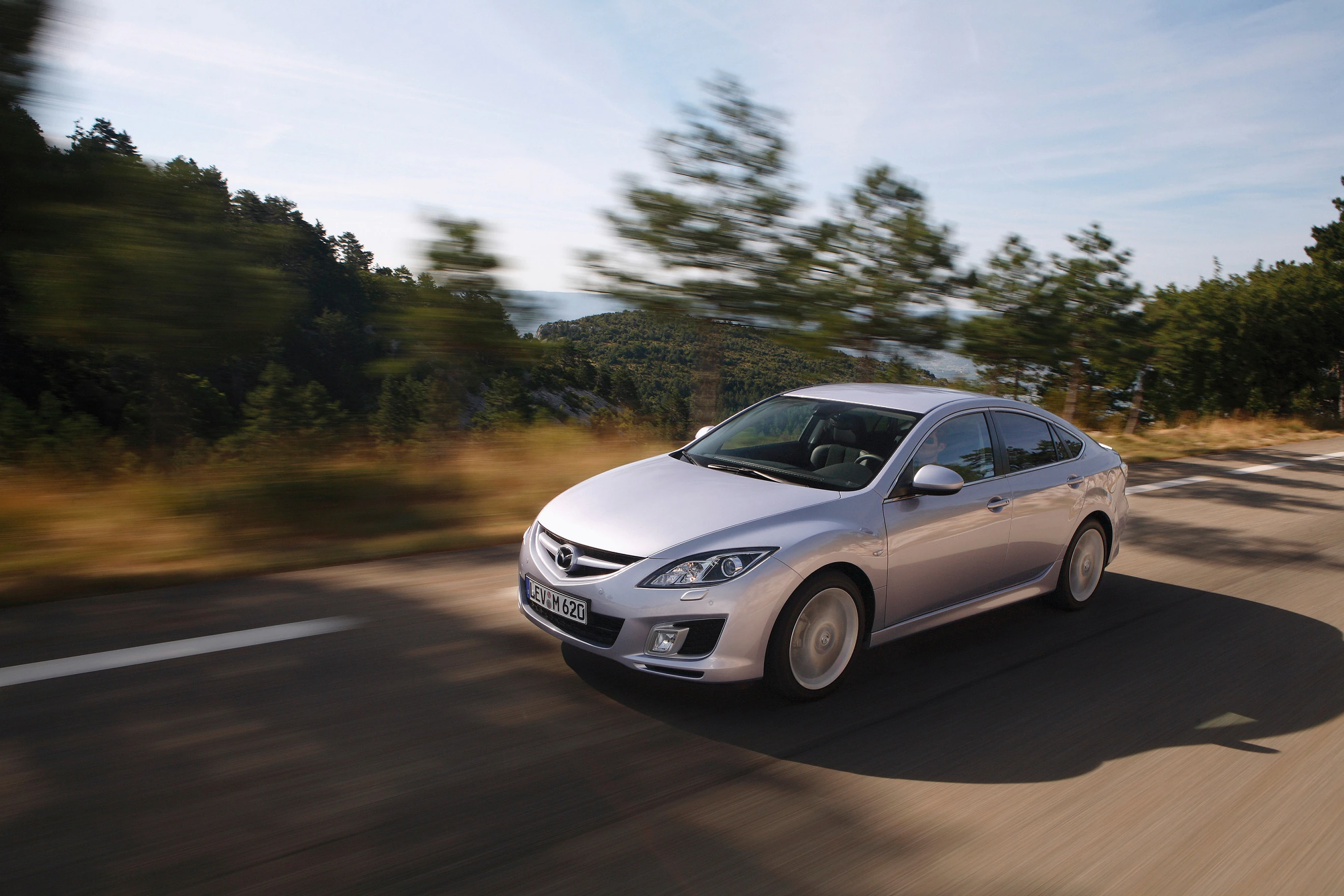 Silberner Mazda 6 fährt auf einer landschaftlichen Straße durch bewaldetes Gebiet bei Sonnenschein. Geschwindigkeit erzeugt Bewegungseffekt.