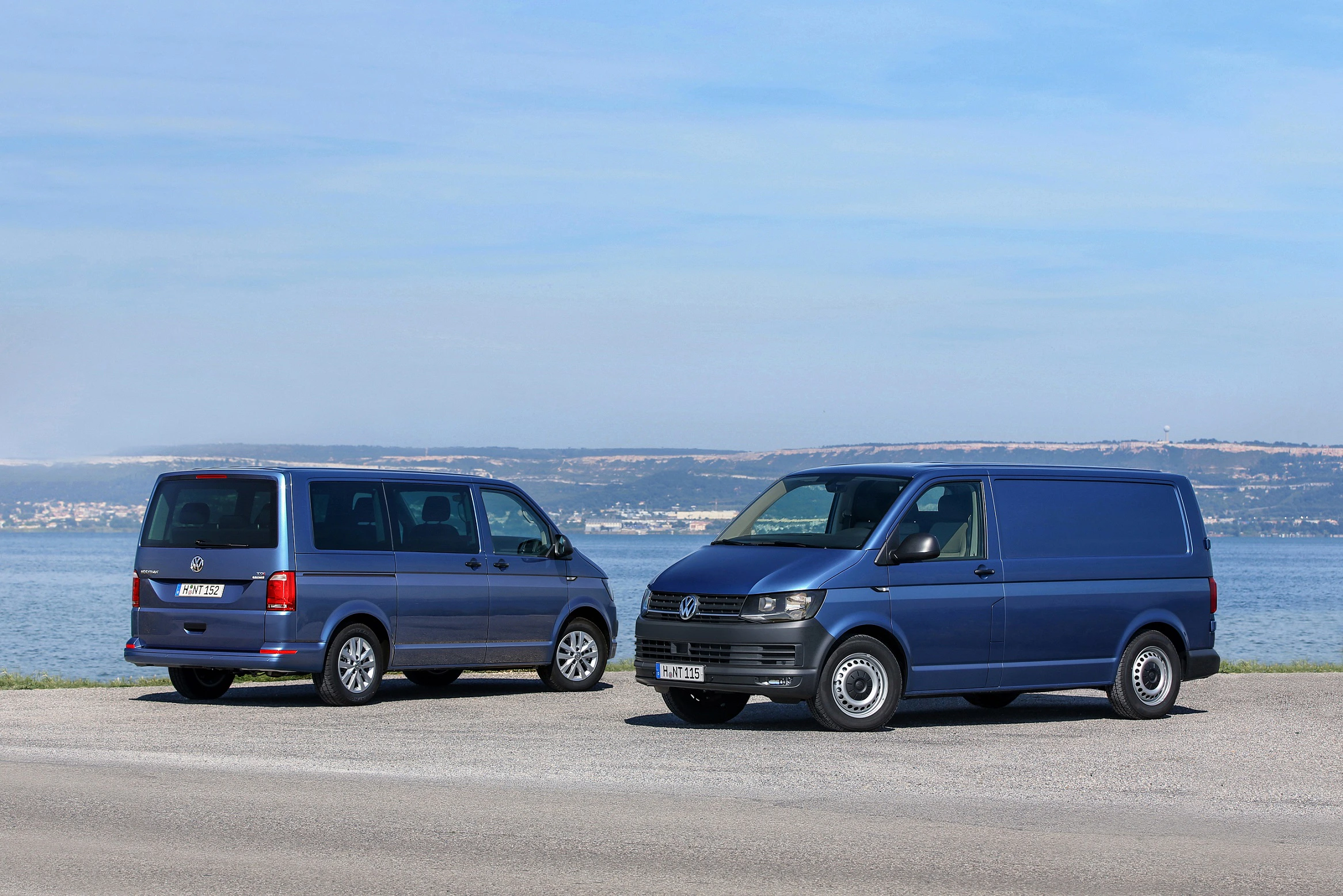 Zwei blaue VW Transporter stehen nebeneinander vor einer Küstenlandschaft mit klarem Himmel und ruhigem Wasser im Hintergrund.