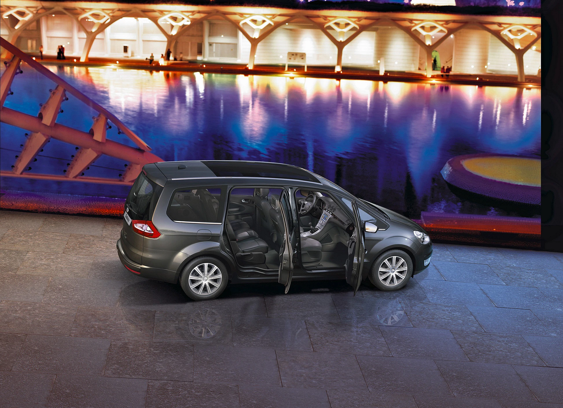 Grauer Minivan mit offenen Türen, steht vor beleuchtetem Gebäude und spiegelndem Wasser, Nachtaufnahme.