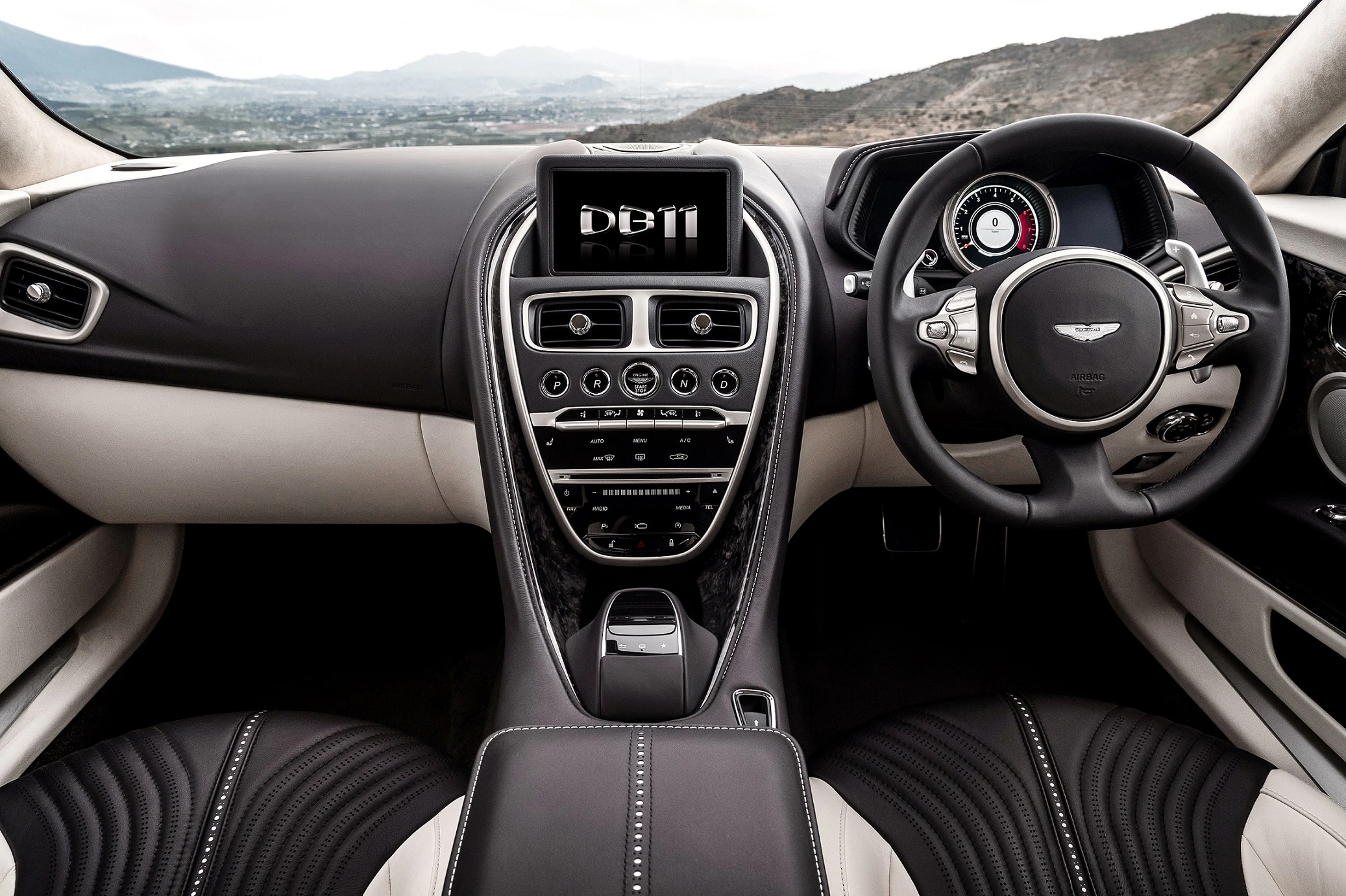Innenraum eines Autos mit schwarzem und weißem Leder, zentralem Bildschirm mit "DB11", Blick auf Lenkrad und Armaturenbrett, Landschaft im Hintergrund.
