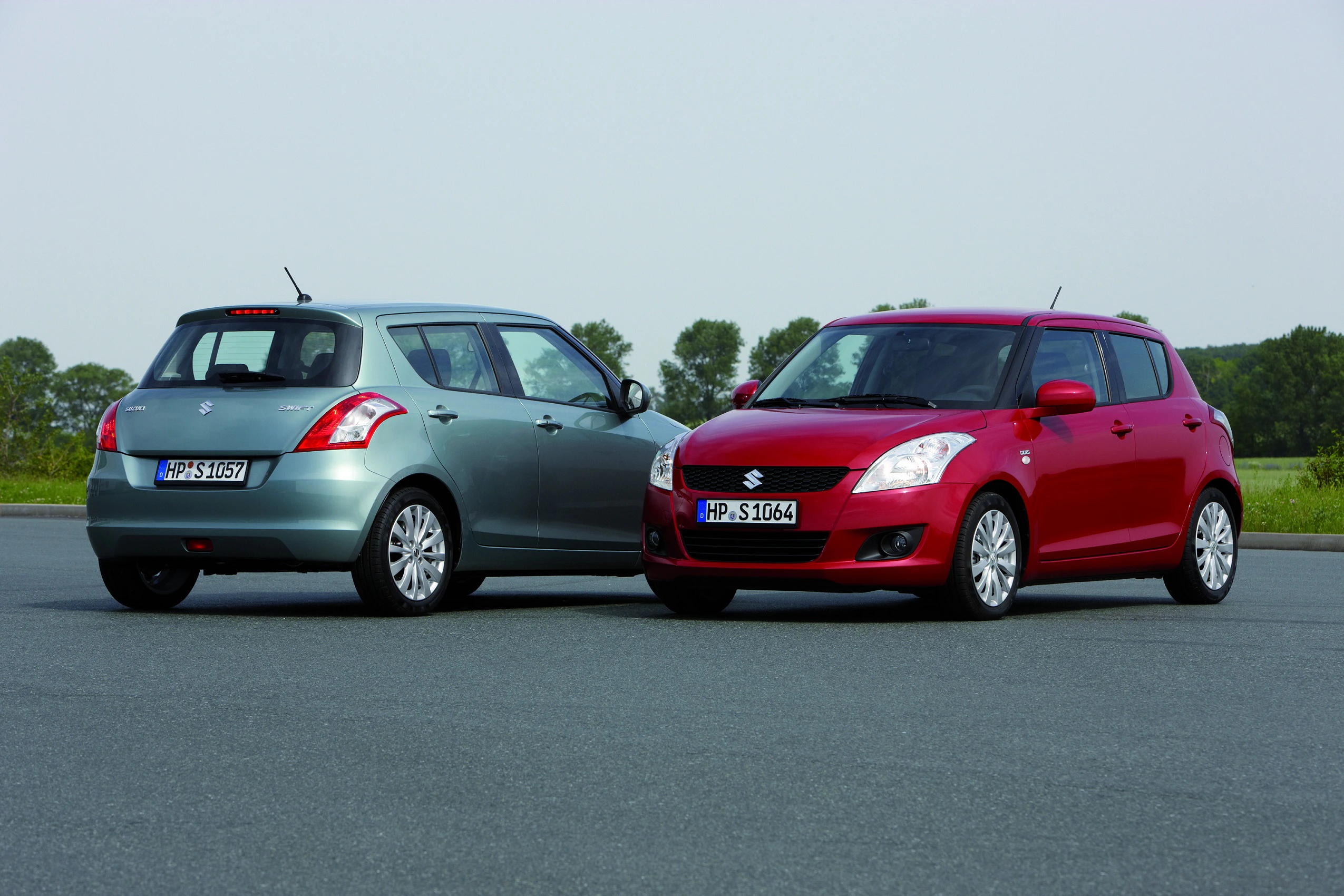 Zwei Suzuki Swift Autos stehen auf einem asphaltierten Platz: ein graues Auto links und ein rotes Auto rechts. Im Hintergrund sind Bäume sichtbar.