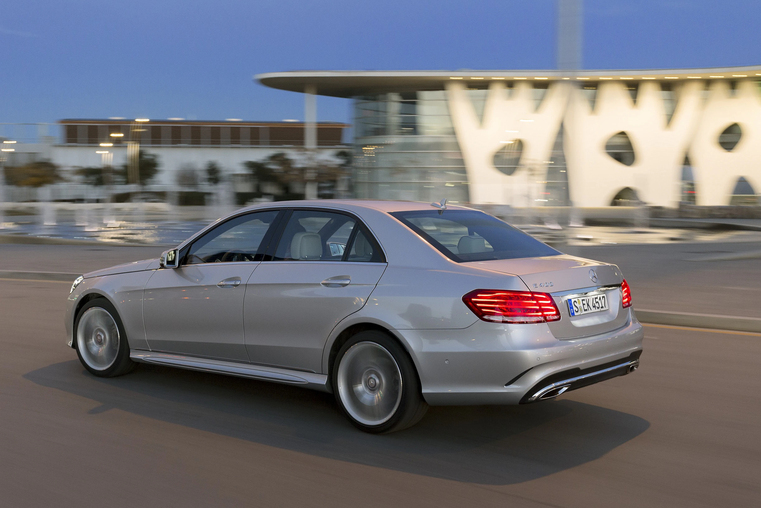 Silbernes Auto fährt bei Dämmerung an einem modernen Gebäude mit organischen Formen vorbei. Dynamische, elegante Limousine.