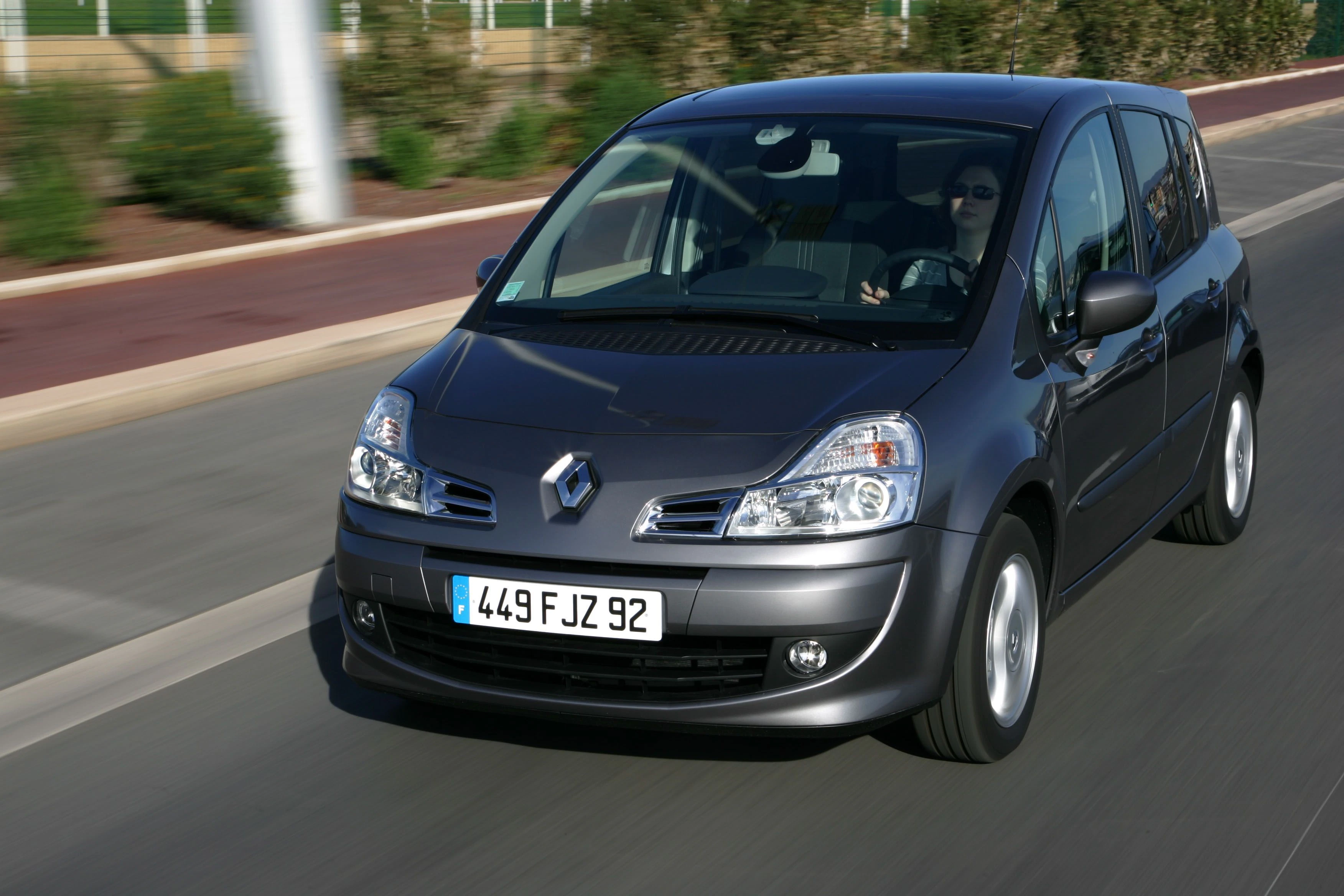 Silberner Renault Scenic fährt auf einer asphaltierten Straße, umgeben von unscharfer Landschaft im Hintergrund.