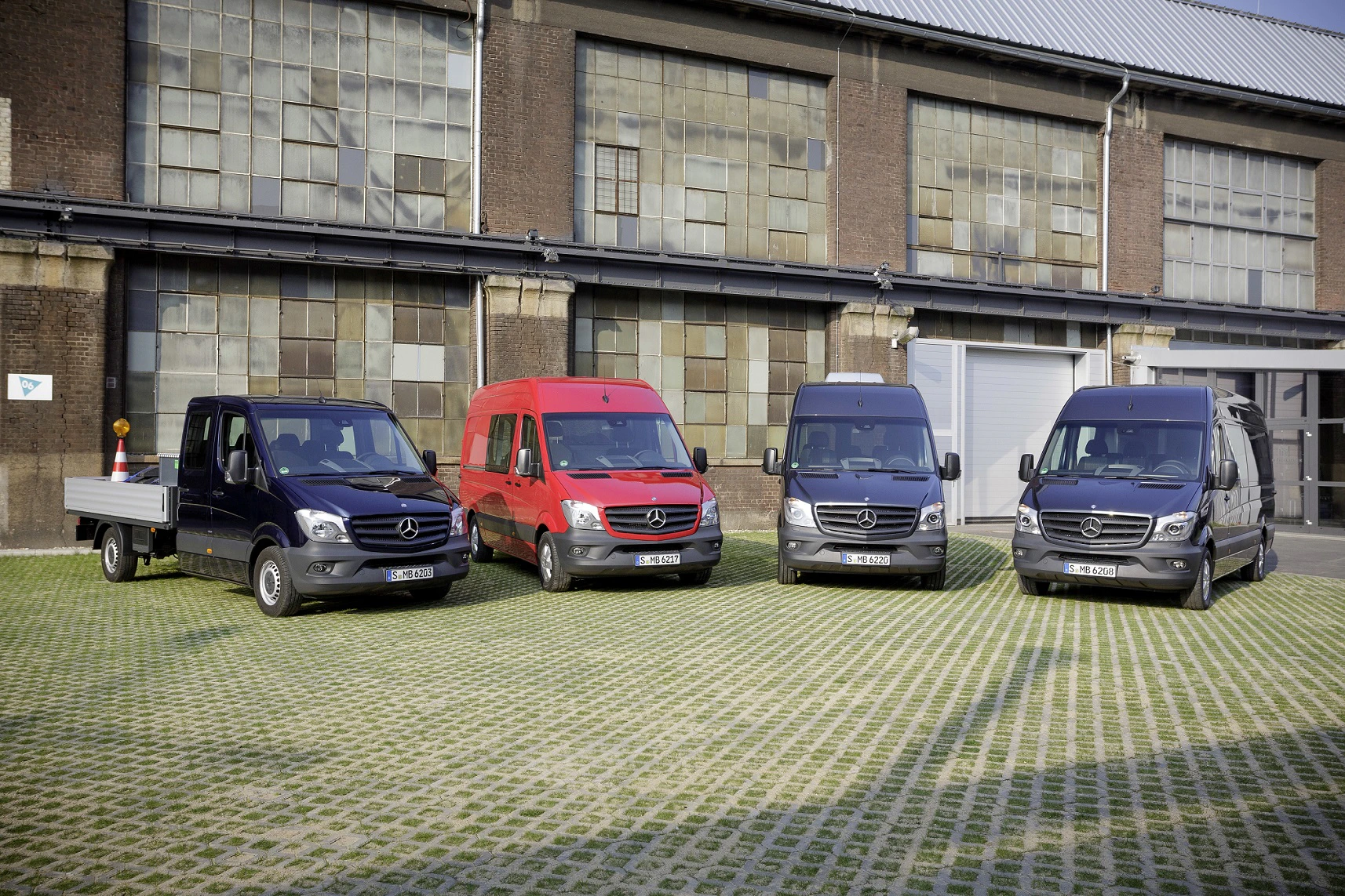 Vier verschiedene Mercedes-Transporter stehen nebeneinander vor einem Industriegebäude mit großen Fenstern und Backsteinwänden.