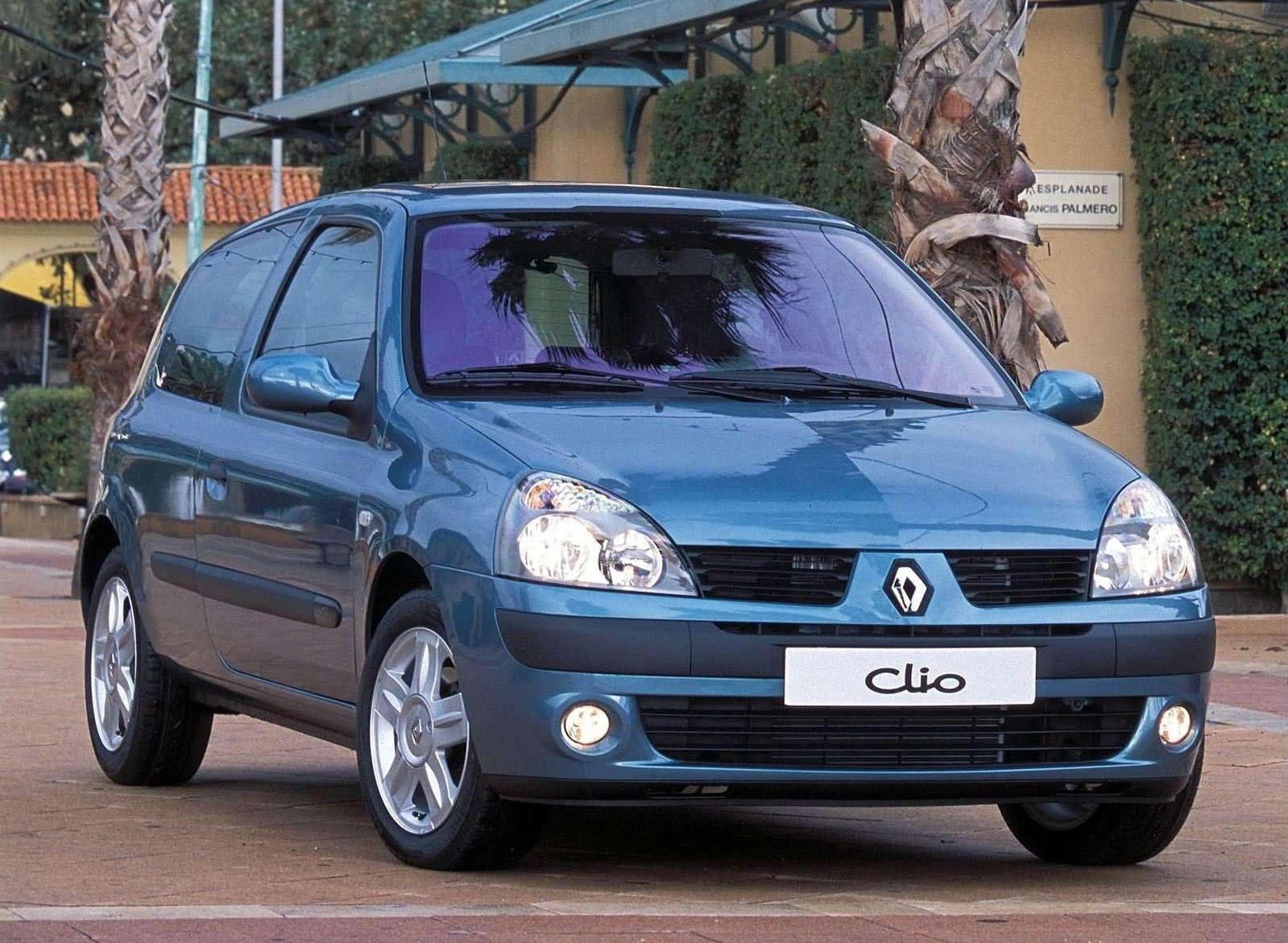 Blauer Renault Clio mit drei Türen steht auf gepflastertem Platz vor Palmen und Gebäuden. Vorderansicht des Fahrzeugs, Scheinwerfer sind an.
