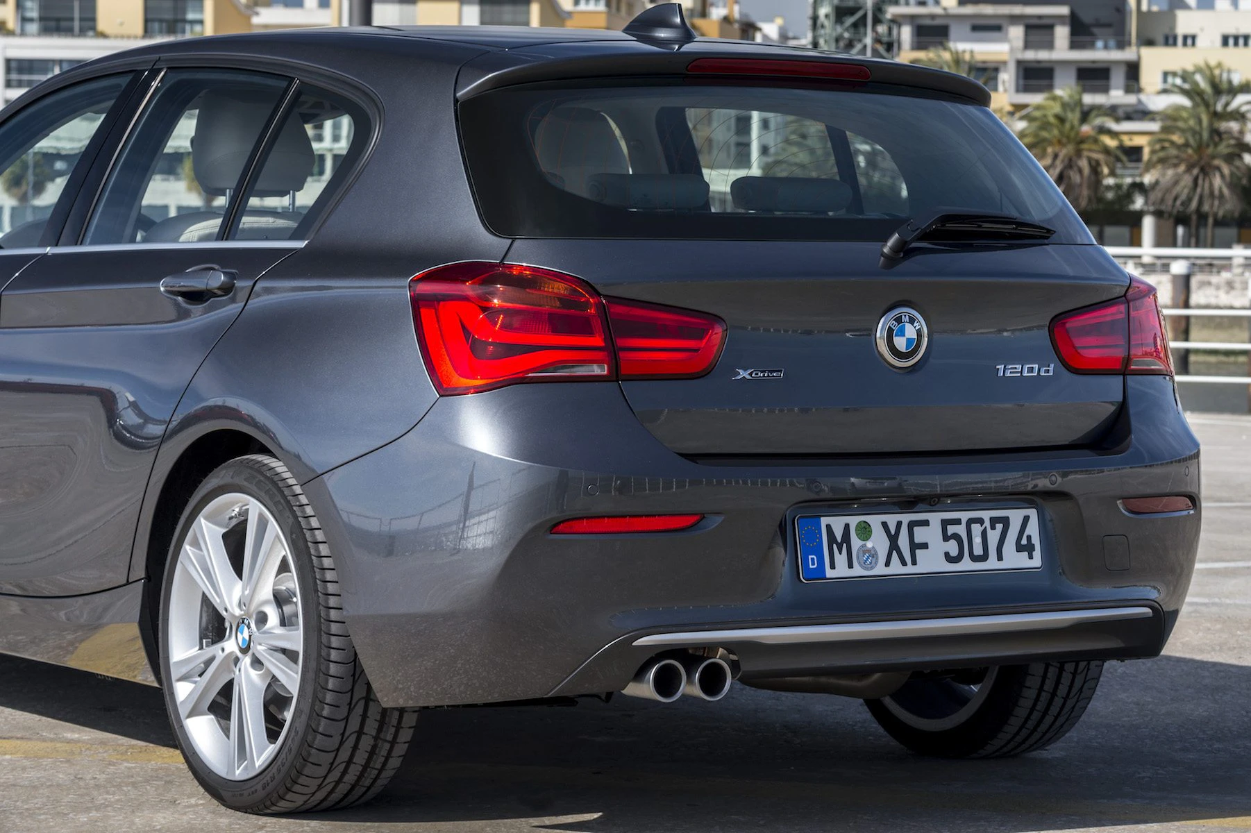 Silberner BMW 120d auf einem Parkplatz, hintere Ansicht. Runde Rückleuchten und Doppelauspuffrohre, Gebäude und Palmen im Hintergrund.