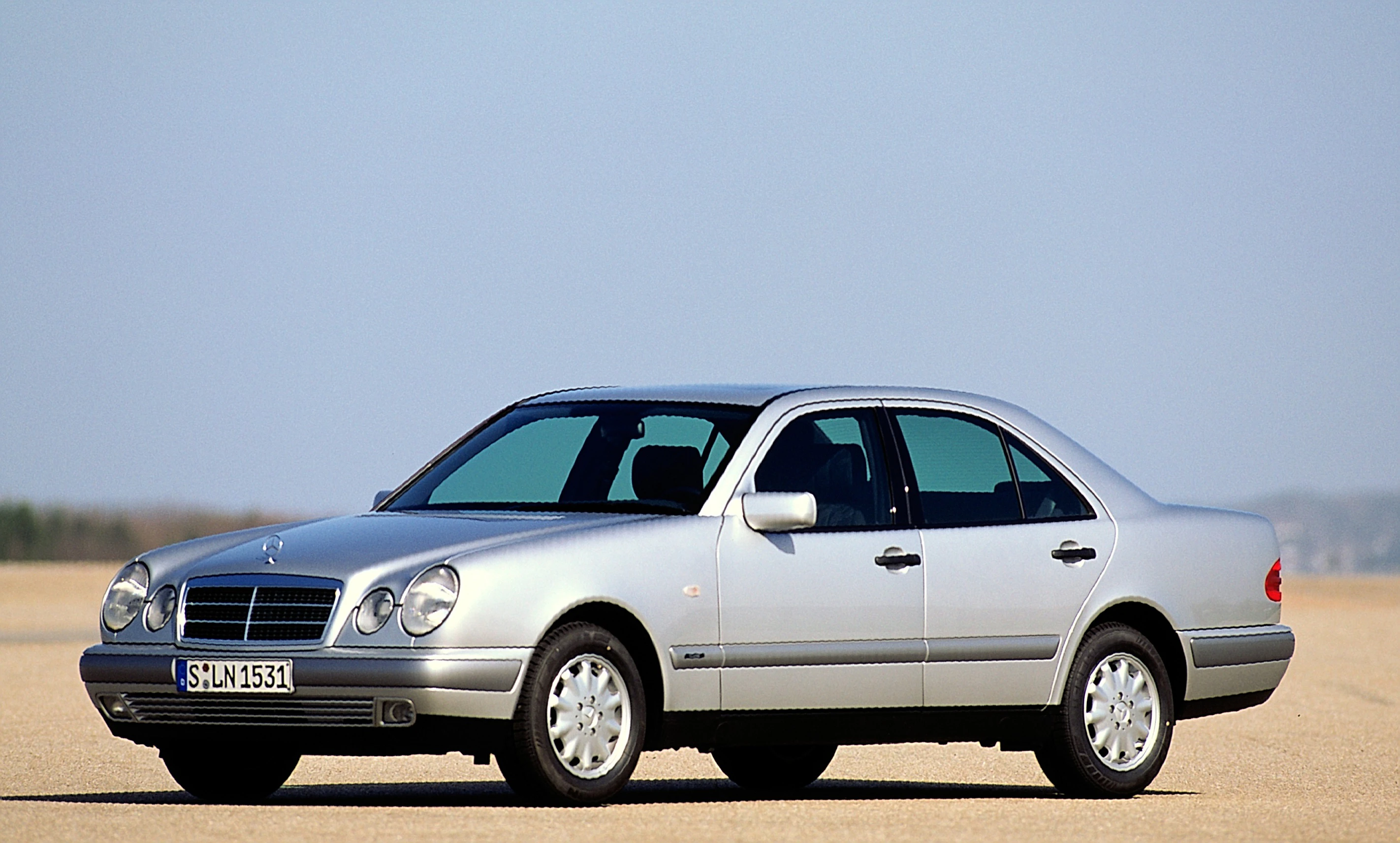Silberner Mercedes-Benz Limousine parkt in einer weiten, sandigen Landschaft bei klarem Himmel. Frontansicht, geradliniges Design.