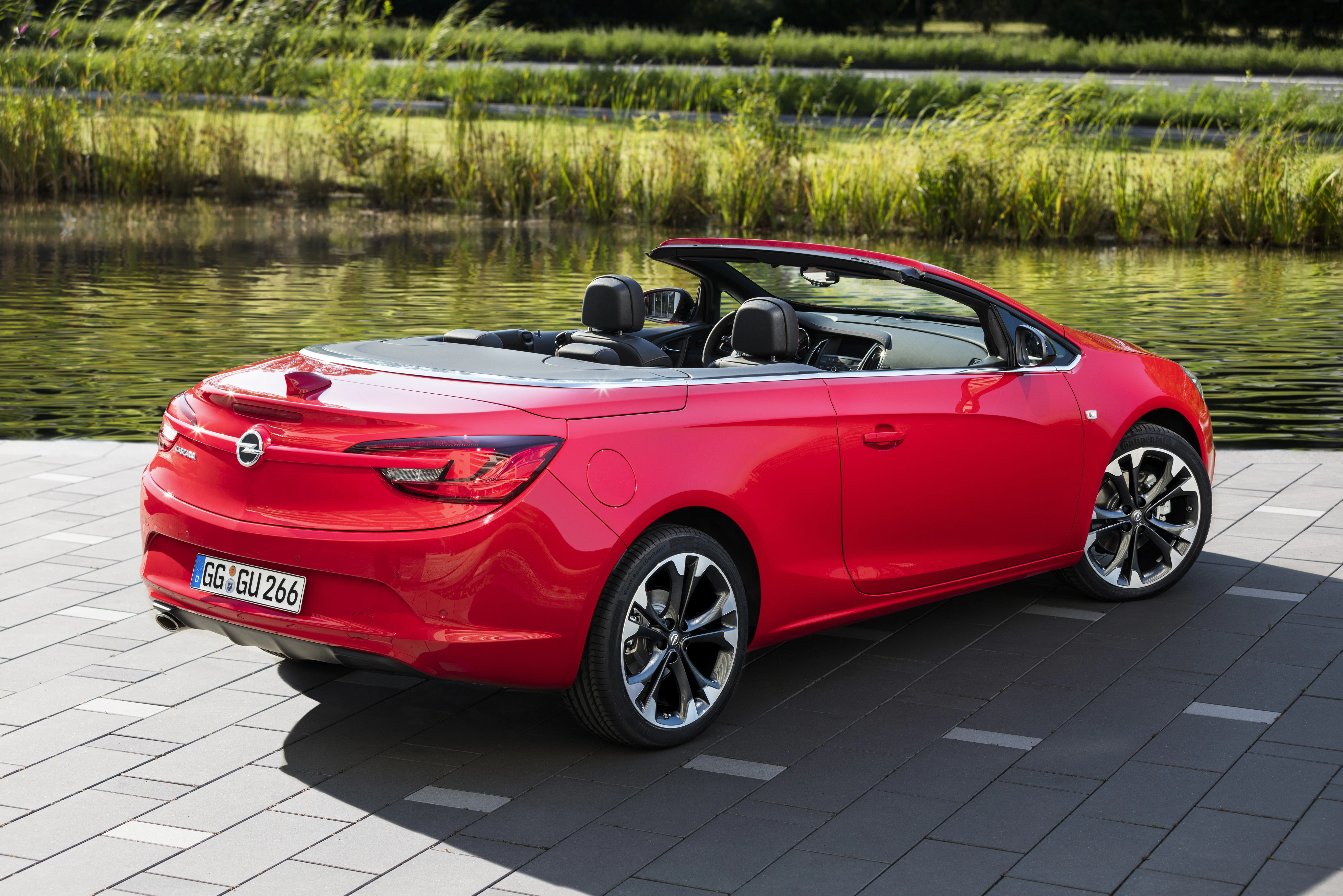 Rotes Cabrio, geparkt auf einer gepflasterten Fläche vor einem Teich. Offen, mit schwarzem Interieur, umgeben von grüner Vegetation.